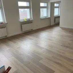 Jasne, przestronne wnętrze po remoncie z nową podłogą z paneli ułożonych w jodełkę, świeżo pomalowane ściany, białe okna z widokiem na budynki za oknem i grzejniki pod oknami; w rogu widoczna...