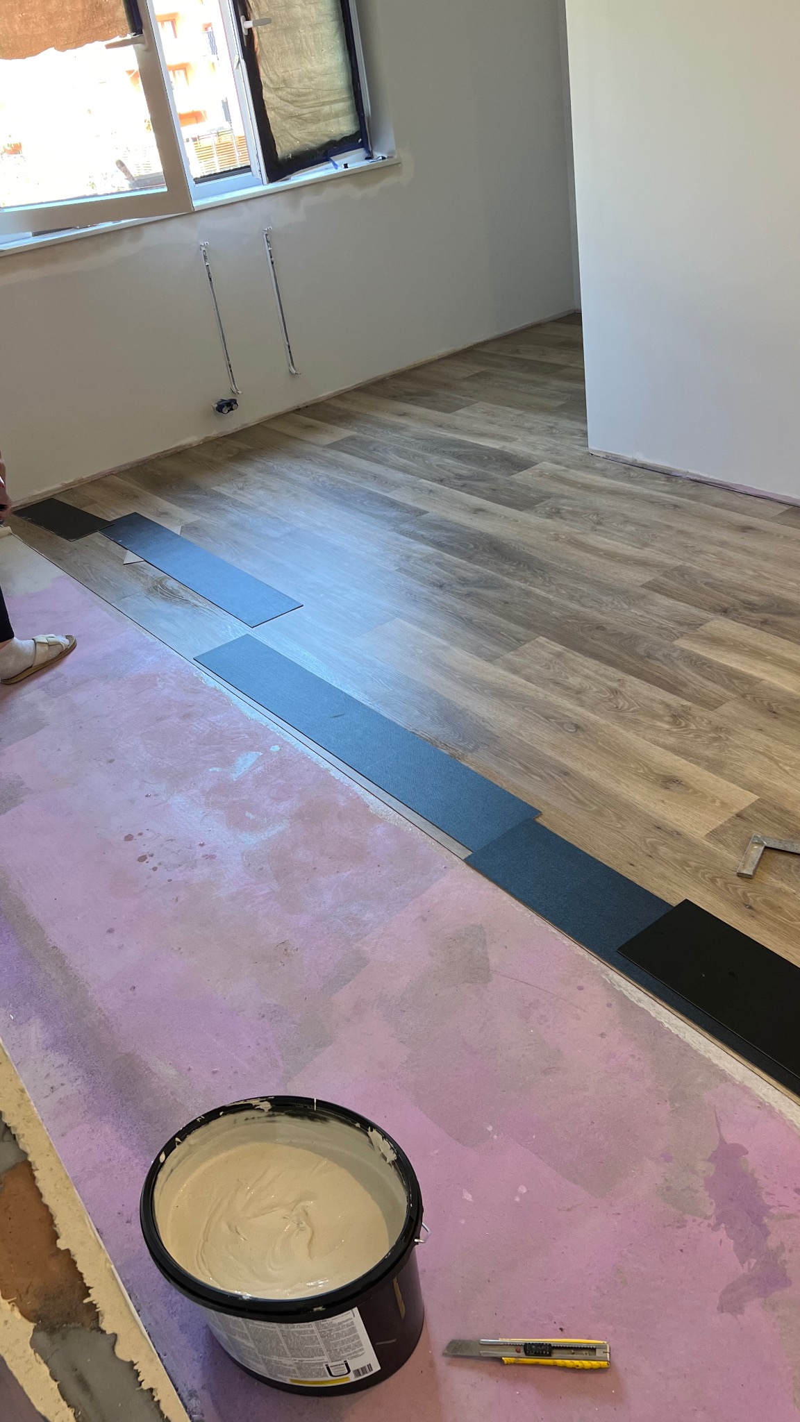 Układanie paneli podłogowych: widoczne panele imitujące drewno, czarne panele w trakcie montażu, wiadro z klejem i nożyk na podłożu z pianki.