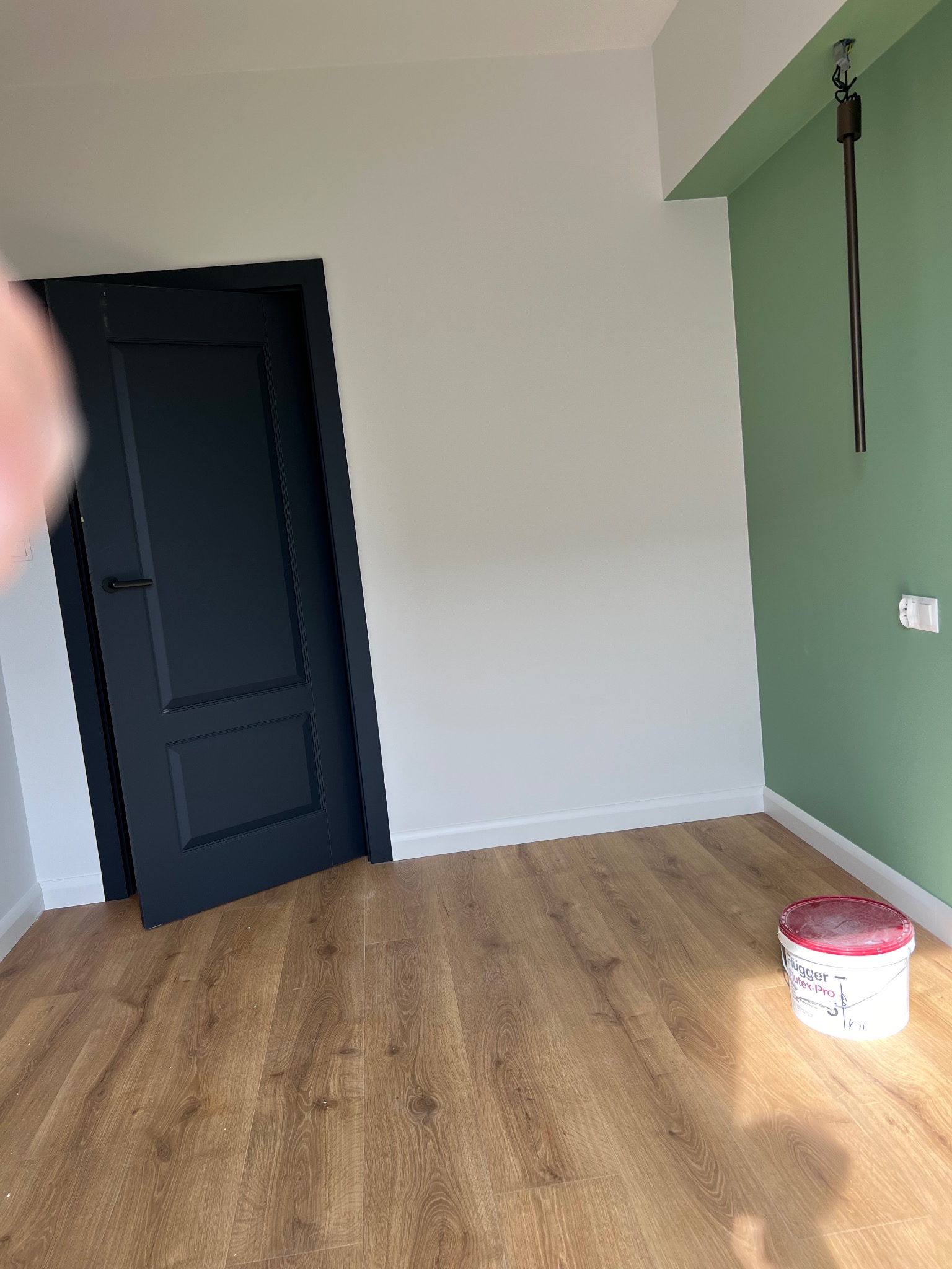 Pomalowane na biało ściany, zielony fragment ściany, ciemne drzwi z czarną klamką, drewnopodobna podłoga i wiadro z farbą Flugger w rogu pokoju.