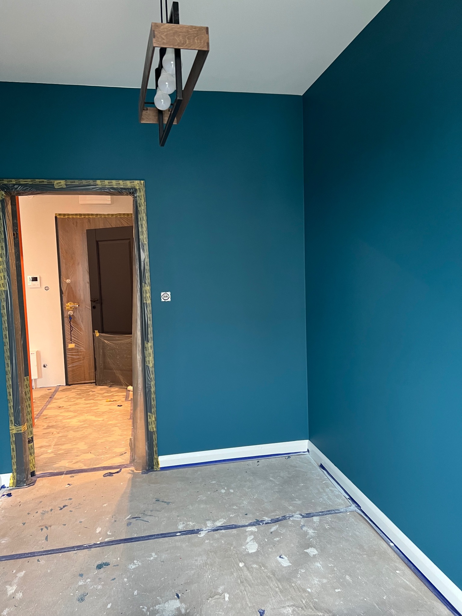 Pokój w trakcie odświeżania: ściany pomalowane na ciemny turkus, podłoga zabezpieczona folią i taśmą malarską, widoczna surowa podłoga, drewniana lampa wisząca w stylu industrialnym, zabezpieczone...