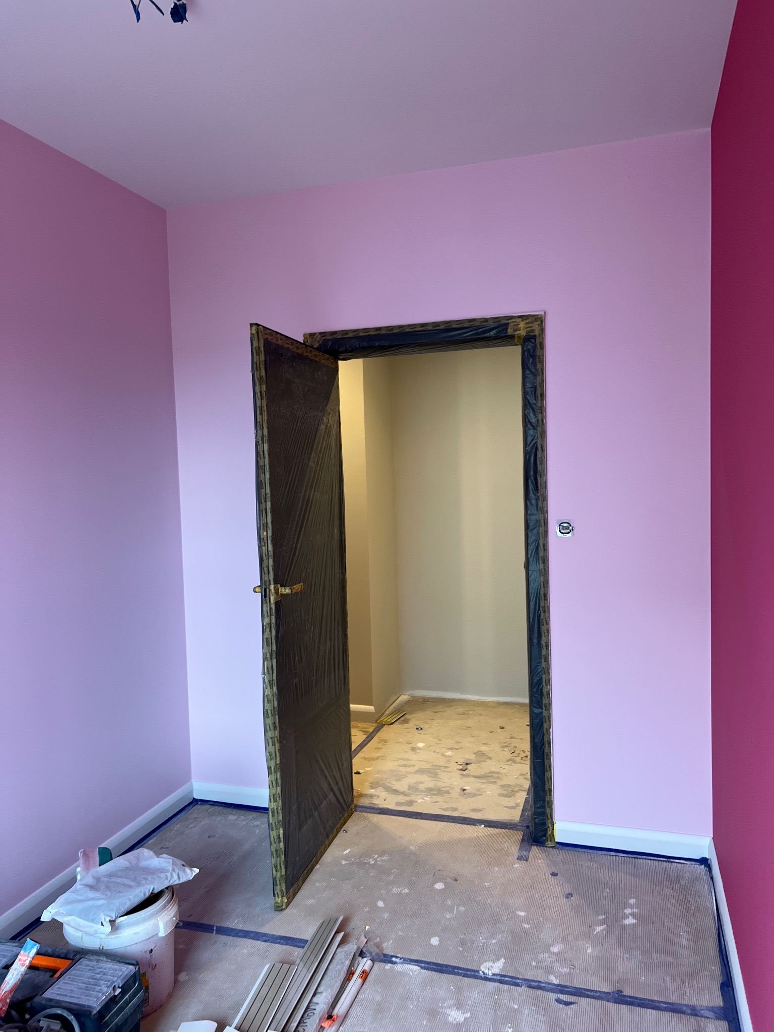 Pokój w trakcie malowania, ściany w odcieniach różu, zabezpieczone folią drzwi, widoczne narzędzia malarskie i listwy przypodłogowe.