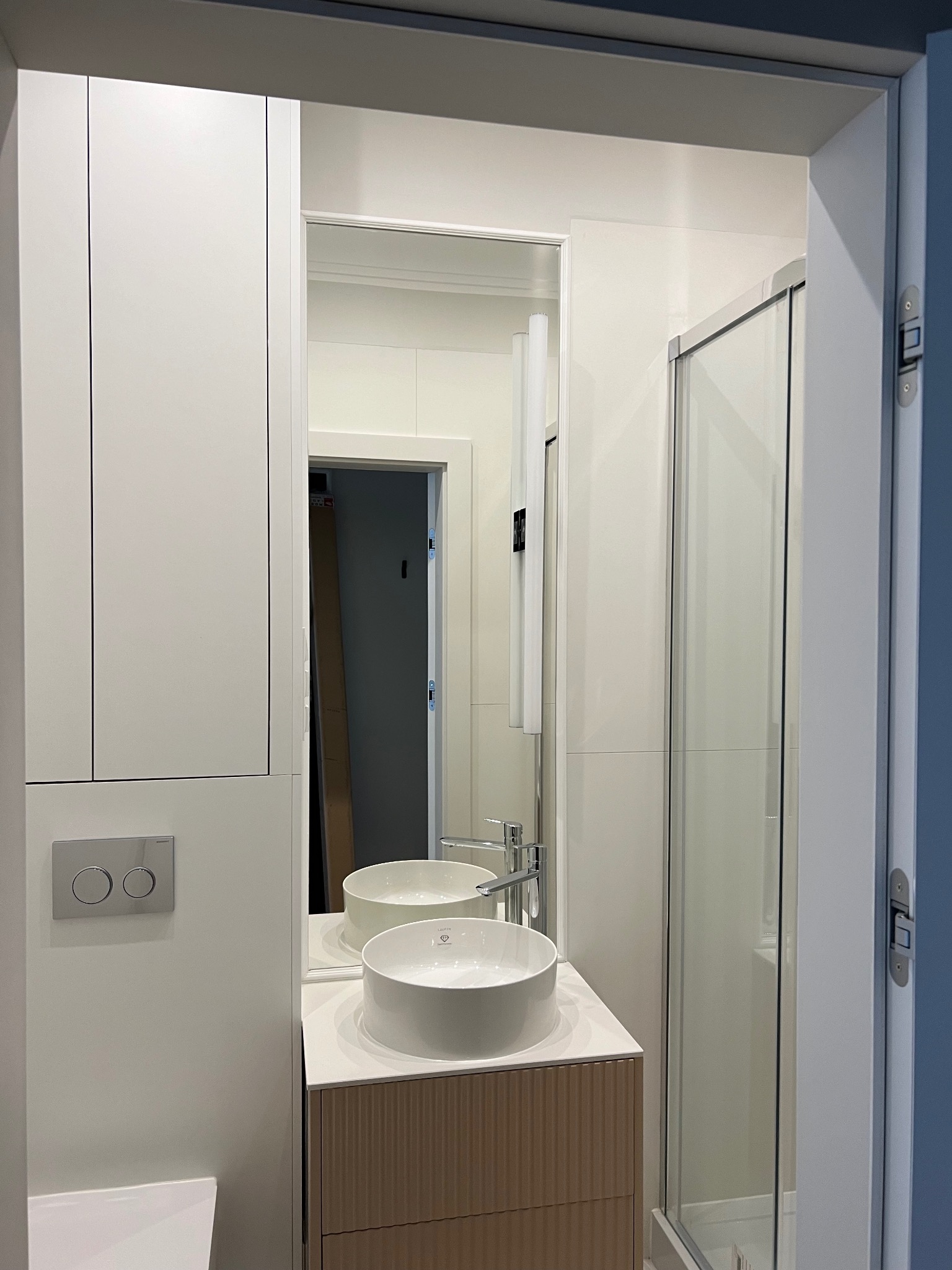 Nowoczesna łazienka z białą umywalką nablatową, szafką z lamelami, kabiną prysznicową i zabudową meblową pod sufit, odbicie lustrzane wnętrza z kartonami.