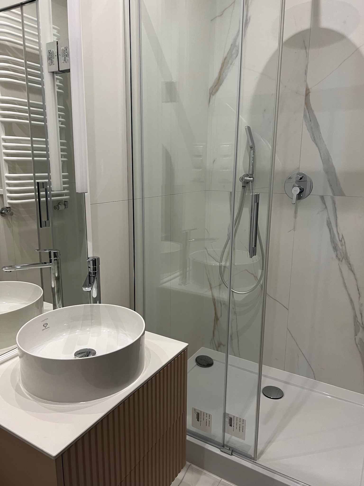Nowoczesna łazienka z kabiną prysznicową ze szklanymi drzwiami, umywalką nablatową marki Laufen na szafce z ryflowanym frontem oraz grzejnikiem drabinkowym w tle.