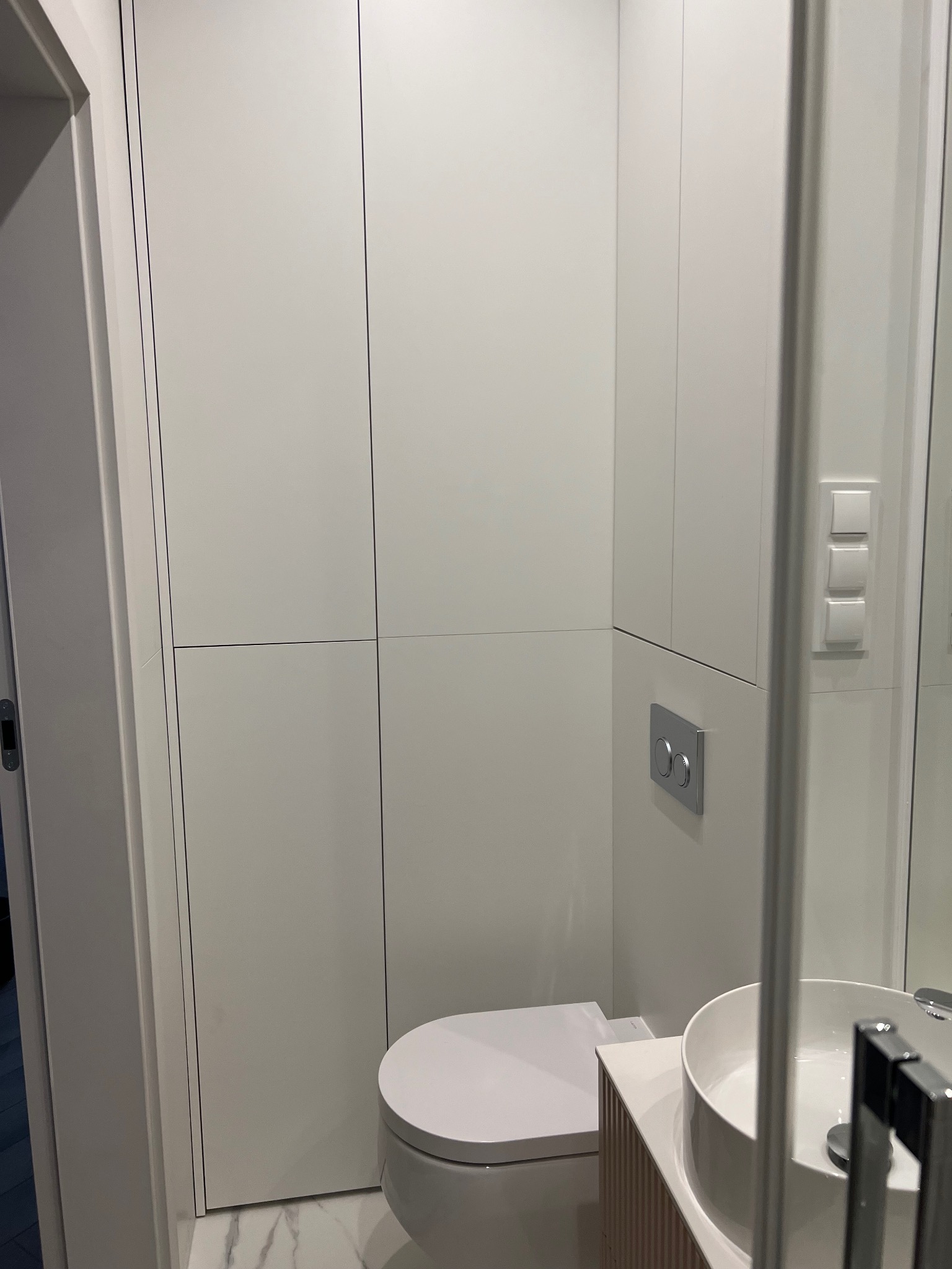 Nowoczesna, minimalistyczna łazienka z białymi szafkami w zabudowie, podwieszaną miską WC i okrągłą umywalką nablatową na szafce z lamelami, marmurowa podłoga.