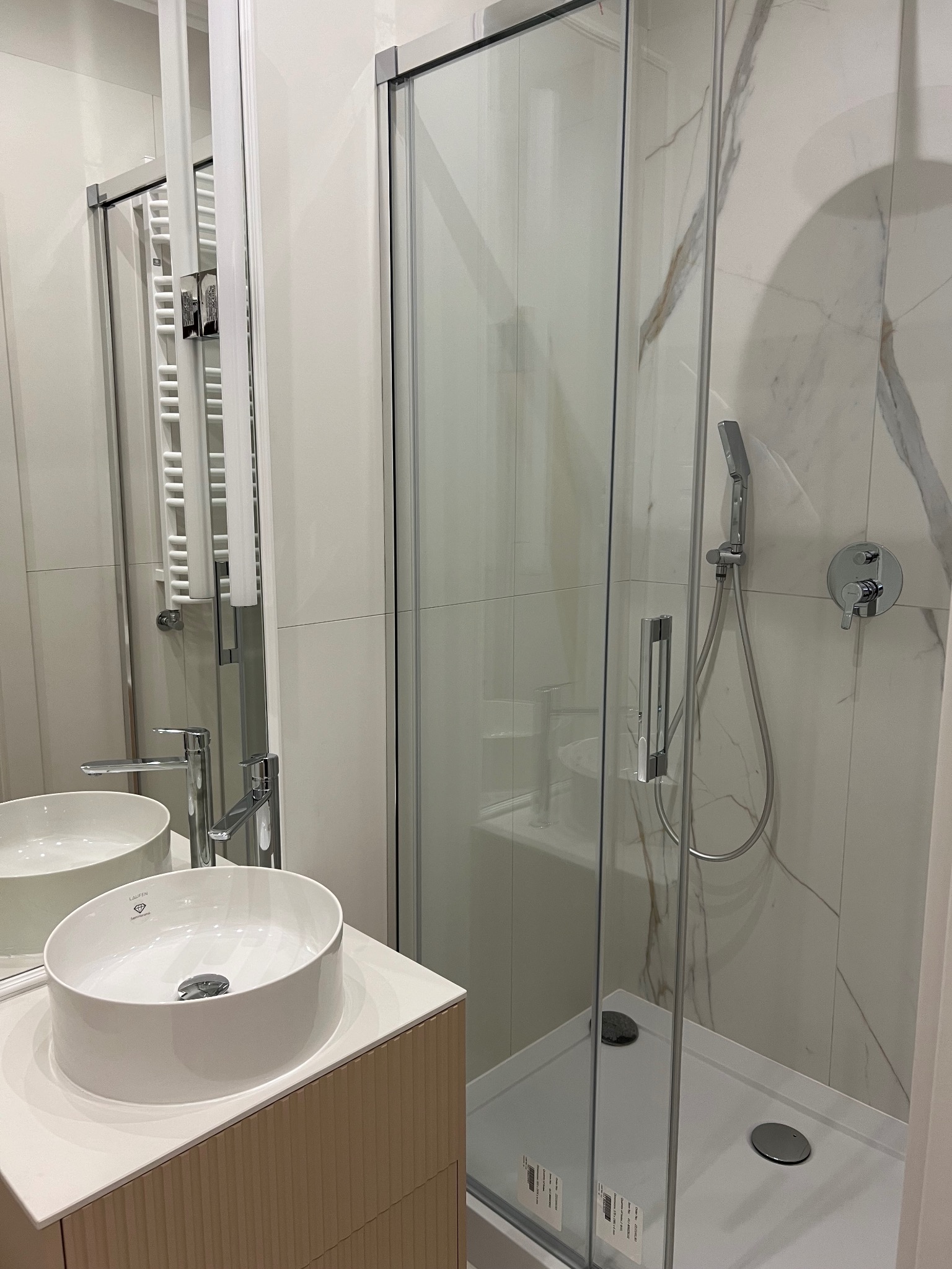 Nowoczesna łazienka z okrągłą umywalką nablatową marki Laufen, kabiną prysznicową ze szklanymi drzwiami i grzejnikiem drabinkowym w jasnej kolorystyce.