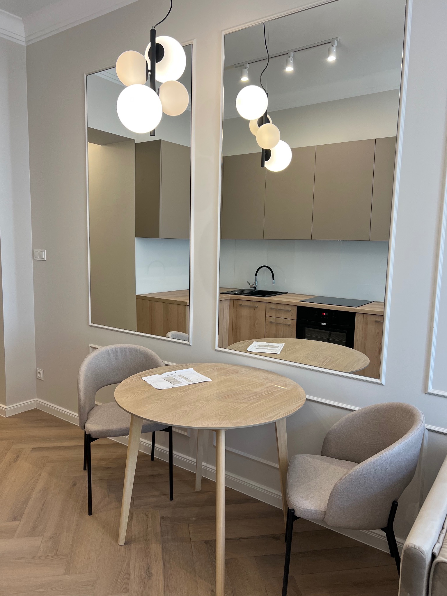 Jasne wnętrze z okrągłym stołem i krzesłami, odbicie kuchni w lustrach na ścianie, wiszące lampy kuliste.