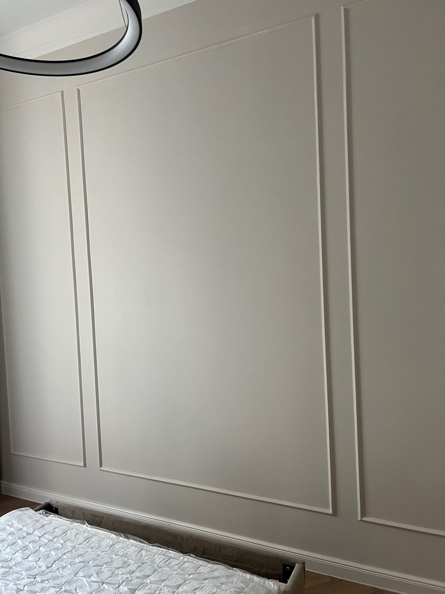 Ściana w pokoju z dekoracyjnymi listwami w stylu klasycznym, pomalowana na jasny odcień szarości, widoczny fragment łóżka z białą narzutą i nowoczesna lampa sufitowa.