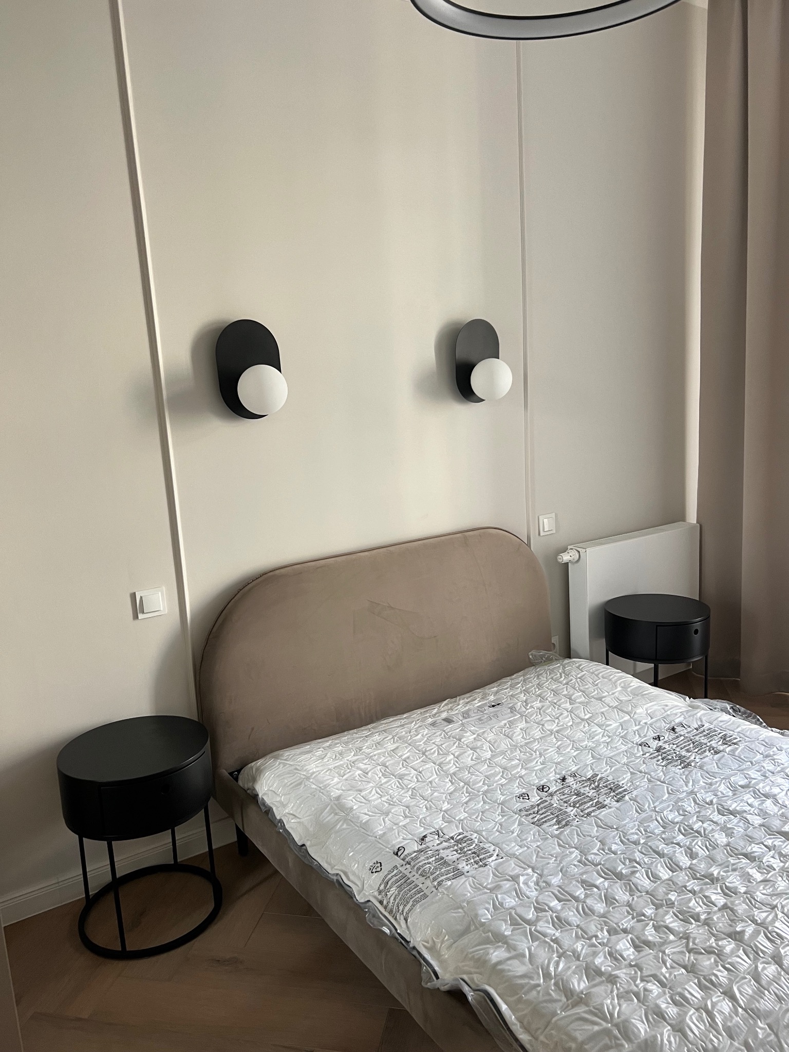 Sypialnia z beżowym zagłówkiem łóżka, czarnymi lampkami nocnymi i okrągłymi stolikami, materac jeszcze w folii ochronnej.