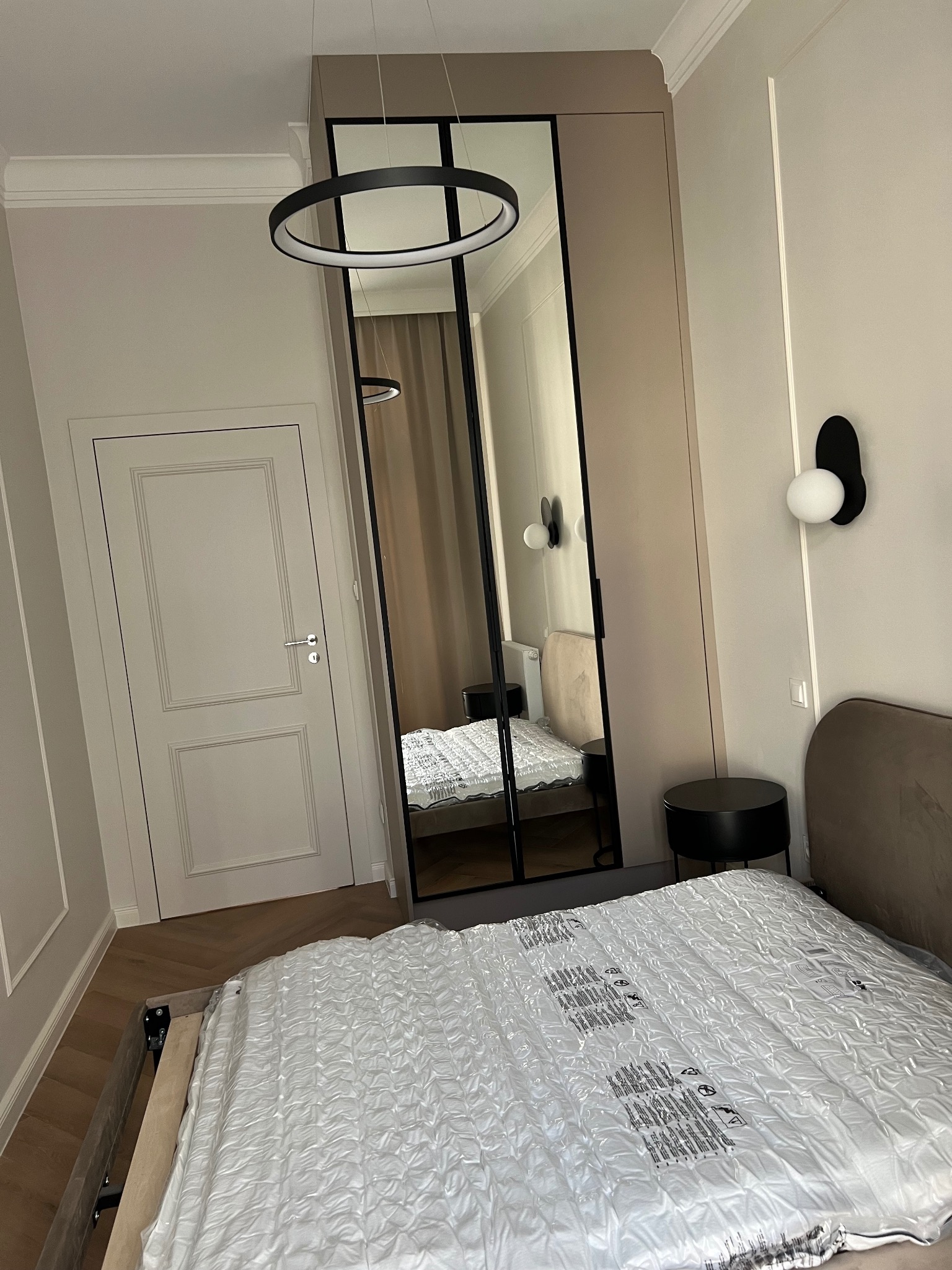 Sypialnia w trakcie urządzania: łóżko z materacem w folii, szafa z lustrem i okrągła lampa sufitowa, widok na drzwi i parkiet.