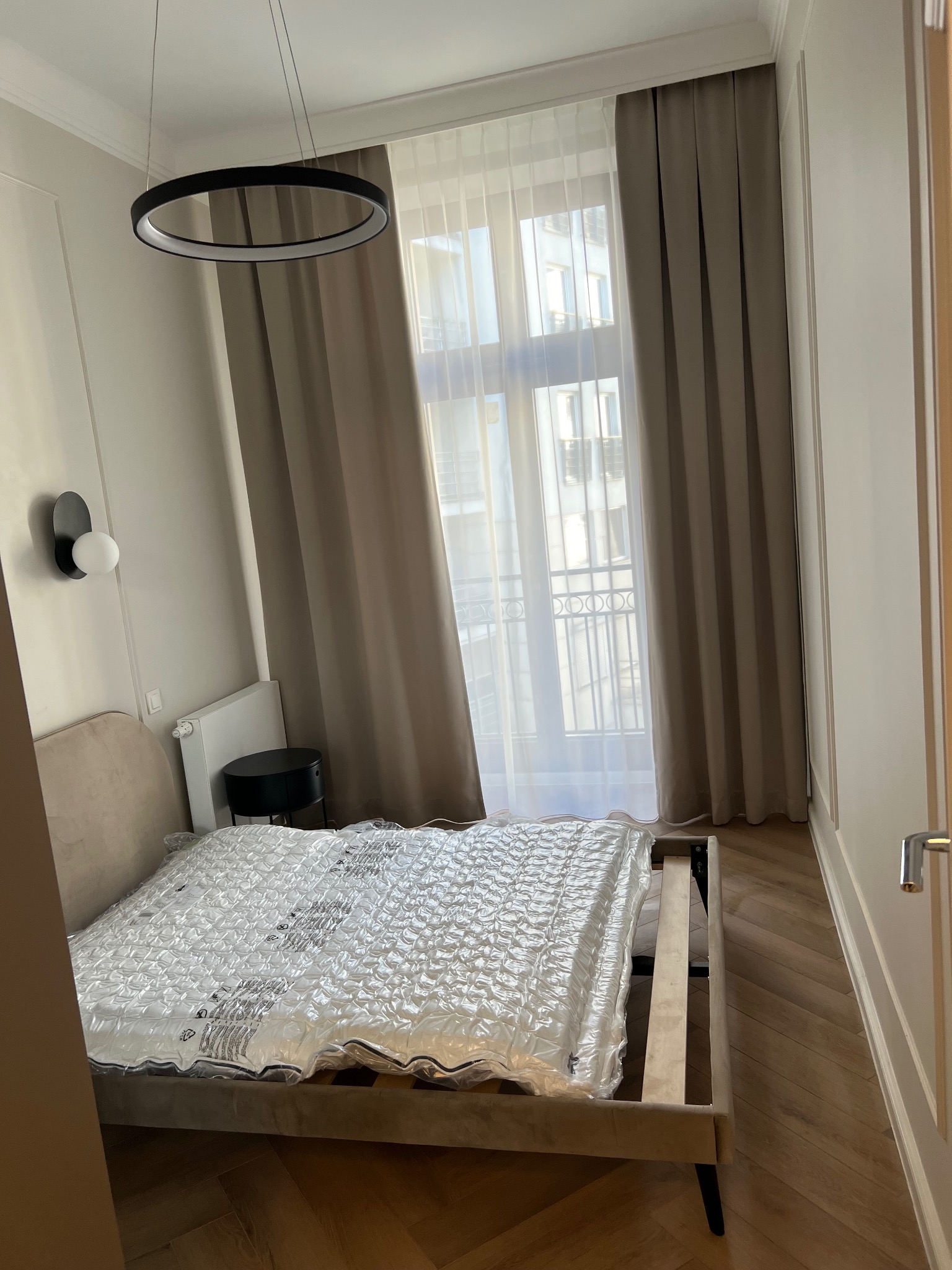 Sypialnia w trakcie wykończenia: łóżko z materacem w folii ochronnej, okrągła lampa sufitowa, beżowe zasłony, parkiet w jodełkę.