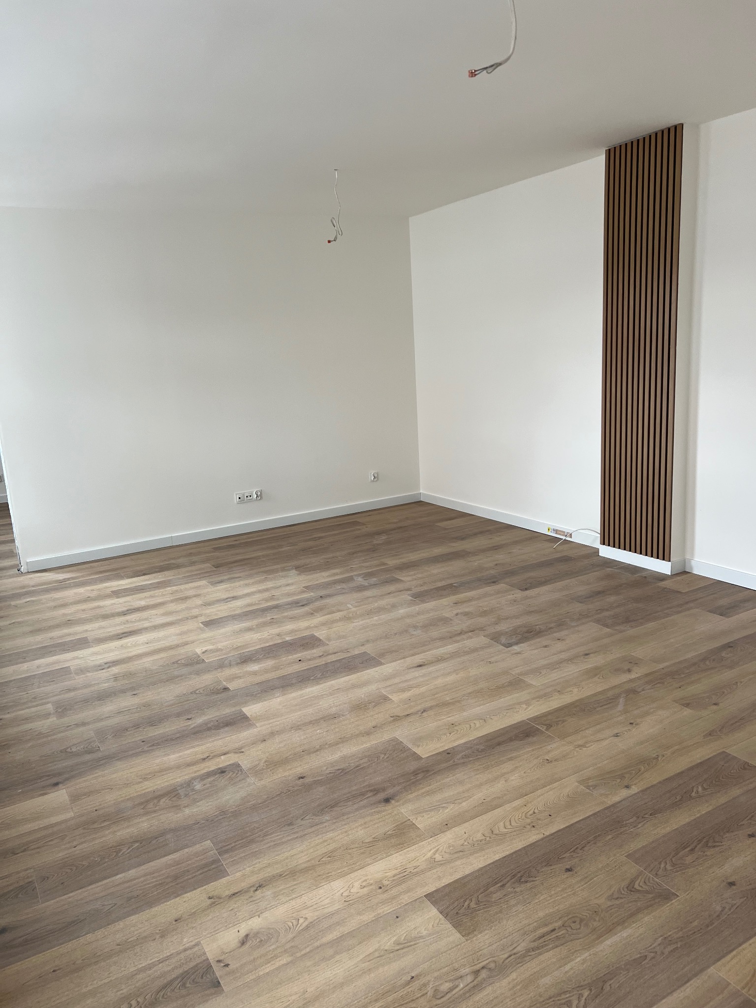 Pusty pokój po remoncie z panelami podłogowymi w odcieniach brązu i pionową dekoracją ścienną z drewnianych listewek; widoczne wystające kable elektryczne.