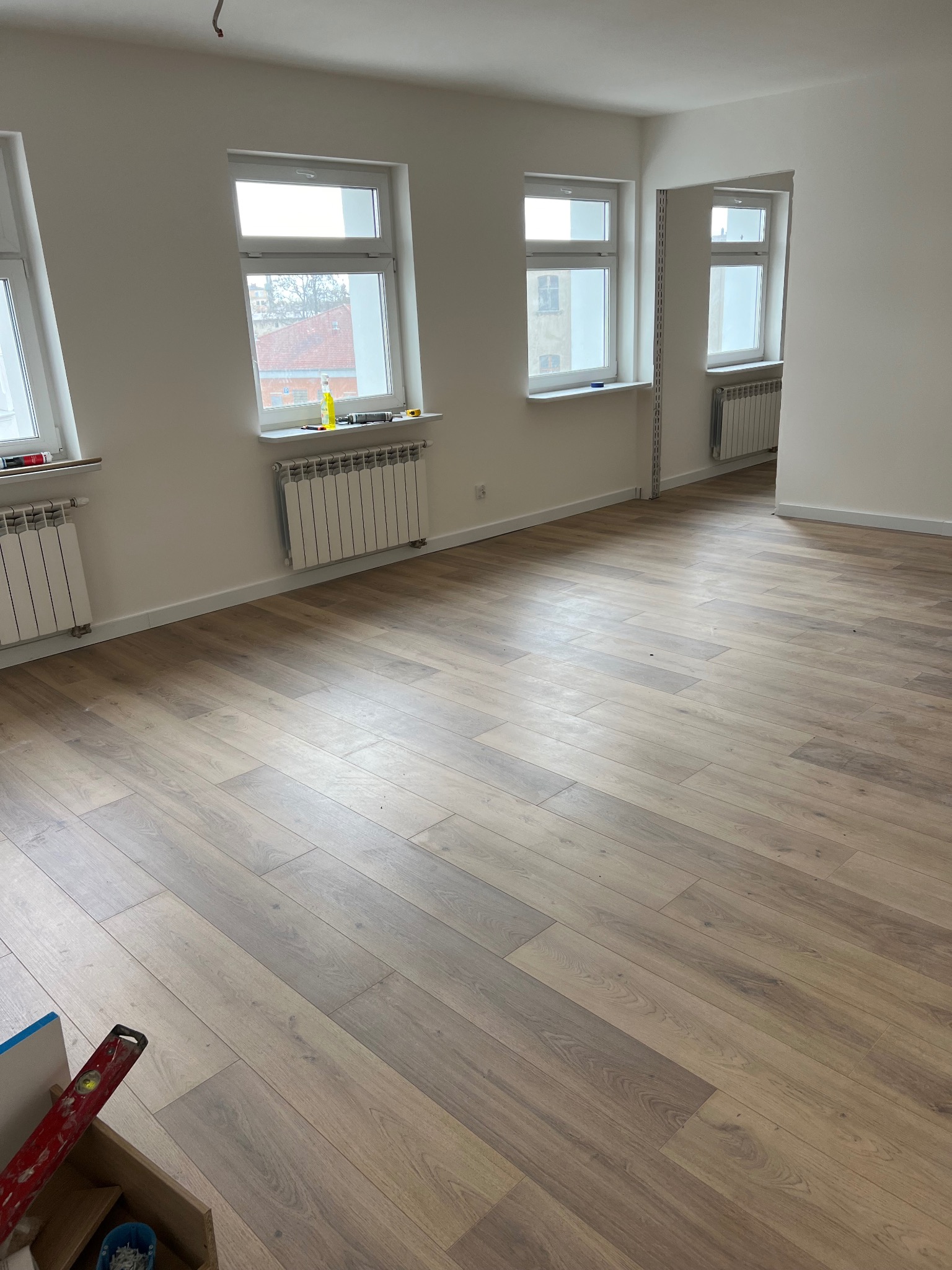 Jasne, przestronne wnętrze po remoncie z nową podłogą z paneli ułożonych w jodełkę, świeżo pomalowane ściany, białe okna z widokiem na budynki za oknem i grzejniki pod oknami; w rogu widoczna...