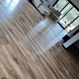 Czysto - Błyszcząca podłoga z paneli imitujących drewno, częściowo widoczny narożnik sofy i okno z widokiem na zieleń.