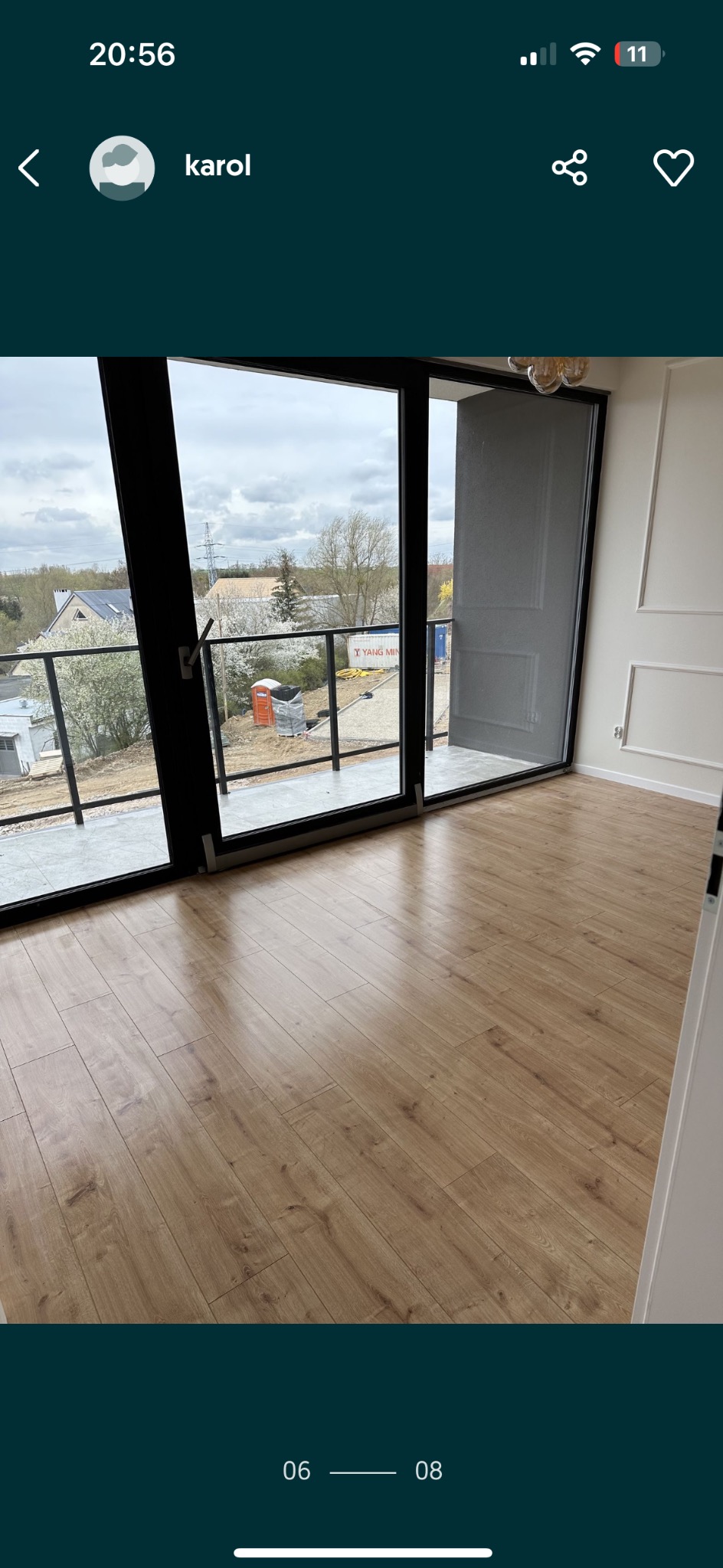 Wnętrze pokoju z drewnianą podłogą, widok na balkon z czarnymi ramami i szklaną balustradą, za którą widać teren budowy z przenośną toaletą i kontenerem.