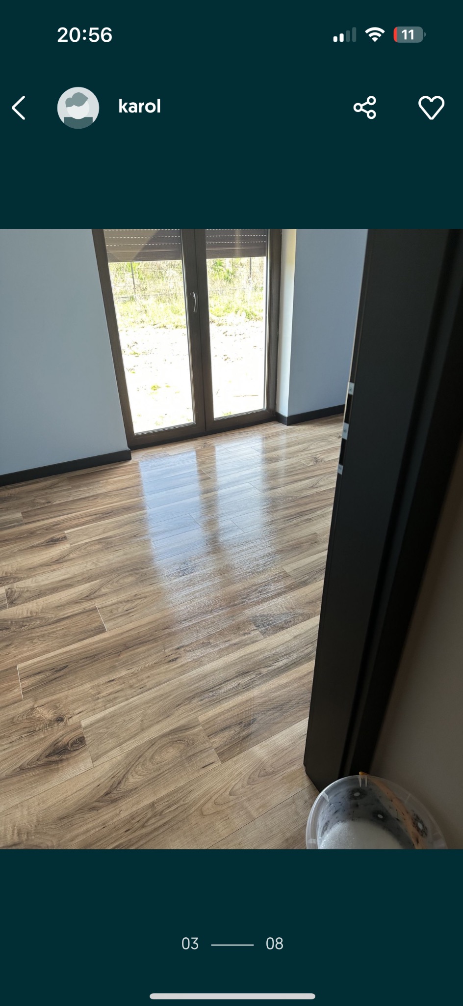 Lśniąca, świeżo umyta podłoga z paneli w pokoju z widokiem na zewnątrz, widoczne wiadro z wodą i detergentem w rogu.