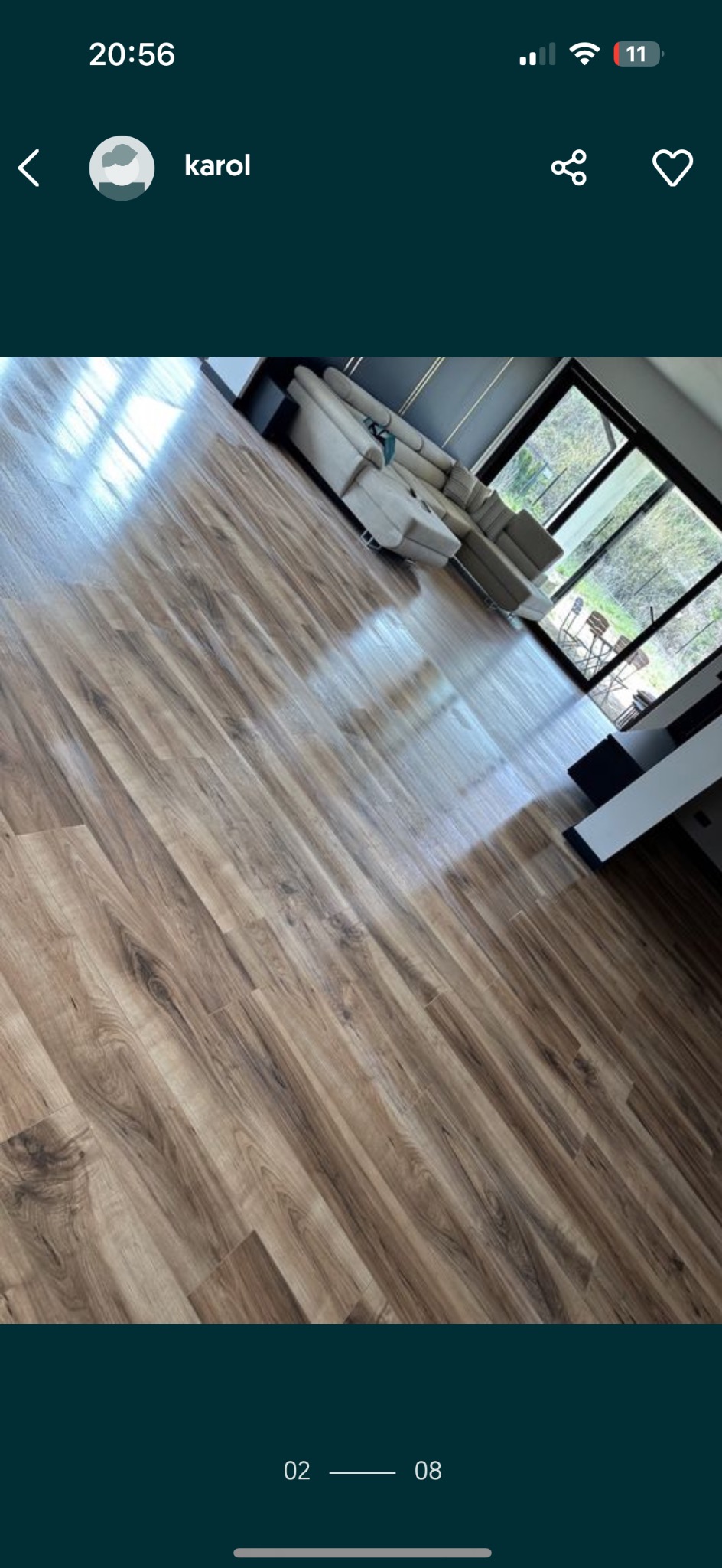 Błyszcząca podłoga z paneli imitujących drewno, częściowo widoczny narożnik sofy i okno z widokiem na zieleń.