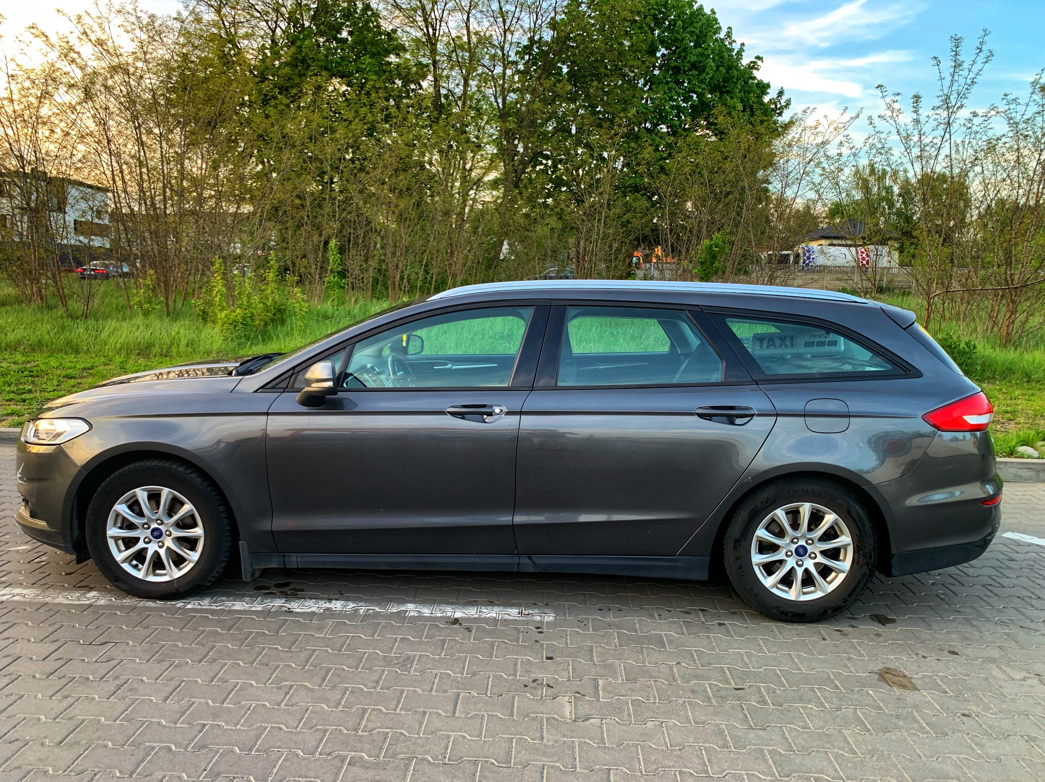 Szary samochód kombi z napisem 'TAXI 800 400 400' na tylnej szybie, zaparkowany na brukowanym parkingu, z zielenią i budynkami w tle.