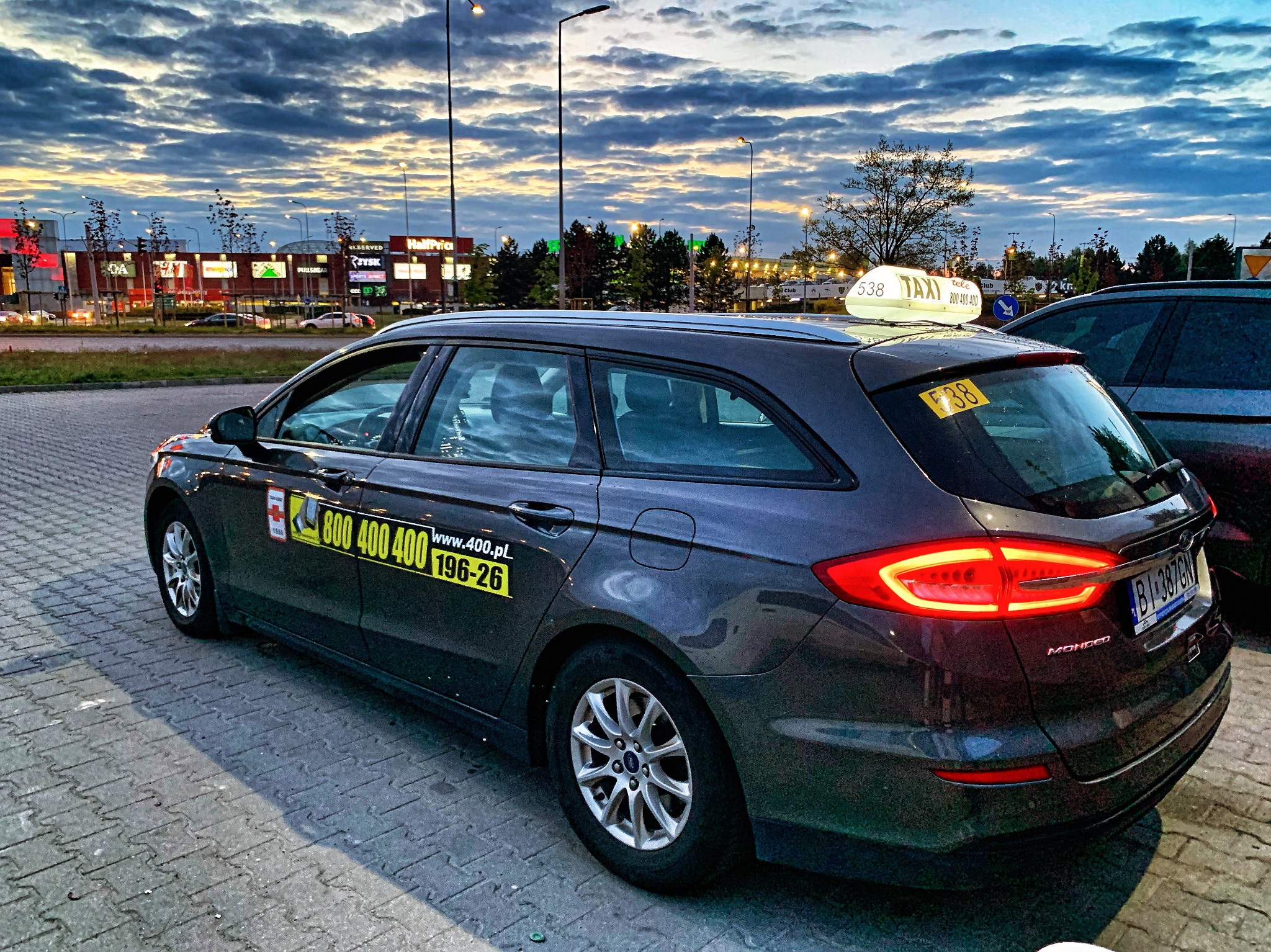 Szary samochód kombi taxi Ford Mondeo z numerem bocznym i lampą taxi na dachu, zaparkowany na kostce brukowej przed budynkiem handlowym o zmierzchu.