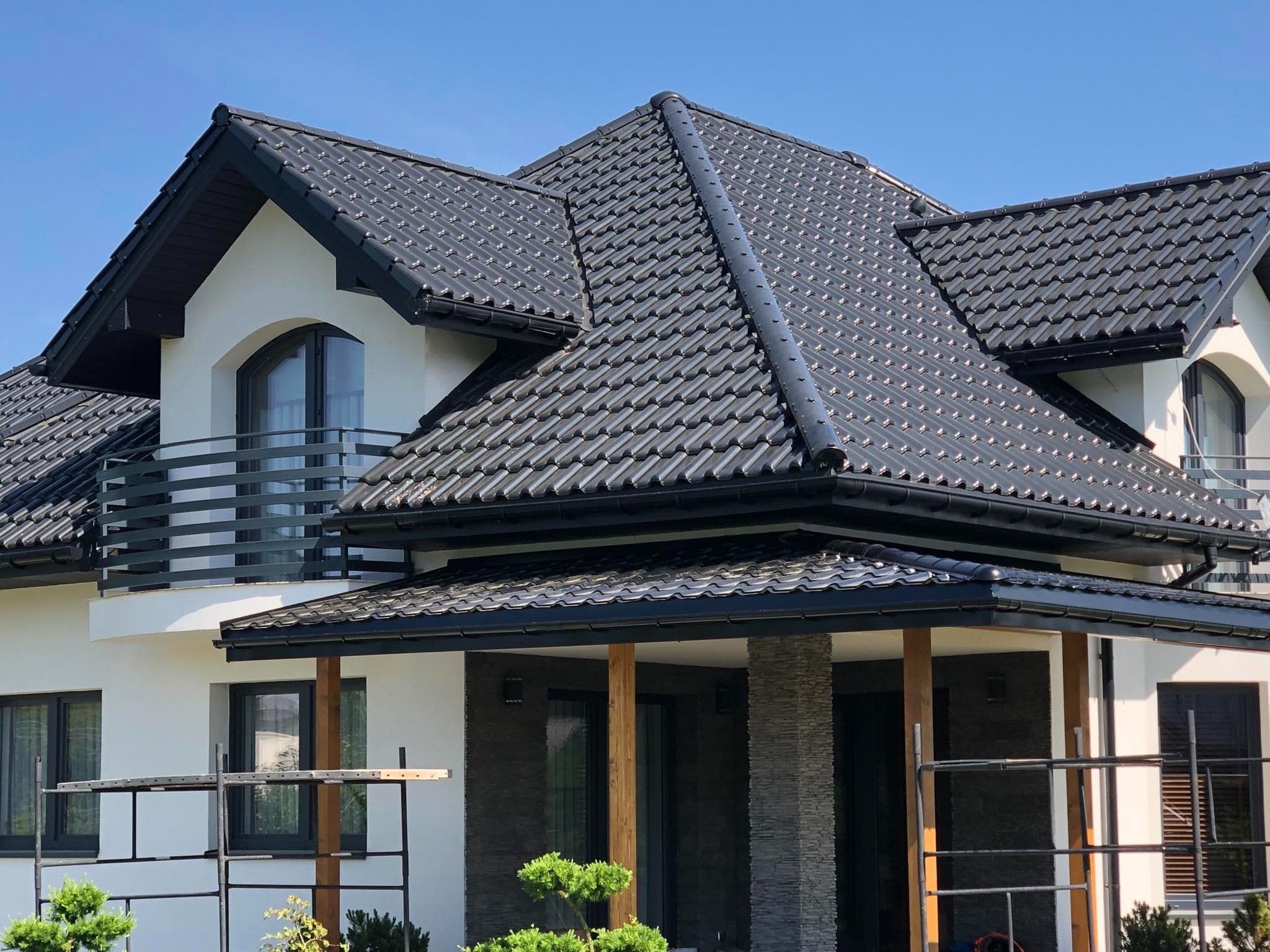 Nowoczesny dom jednorodzinny z ciemnoszarą dachówką ceramiczną, czarnymi rynnami i obróbkami blacharskimi, widoczne rusztowania na pierwszym planie.