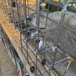 Zbrojenie fundamentu w formie stalowej klatki, połączone drutem wiązałkowym, wsparte na drewnianym szalunku i metalowych podporach, widoczne elementy krajobrazu w tle.