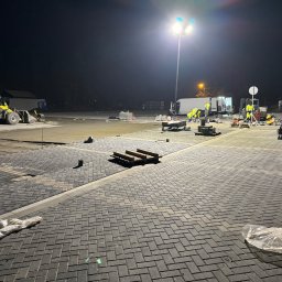 MIKOLO LOGISTIC SPÓŁKA Z OGRANICZONĄ ODPOWIEDZIALNOŚCIĄ - Nocna praca przy układaniu kostki brukowej na dużej powierzchni, widoczny sprzęt budowlany, paleta z kostką, ekipa w odblaskowych ubraniach i oświetlenie placu budowy.