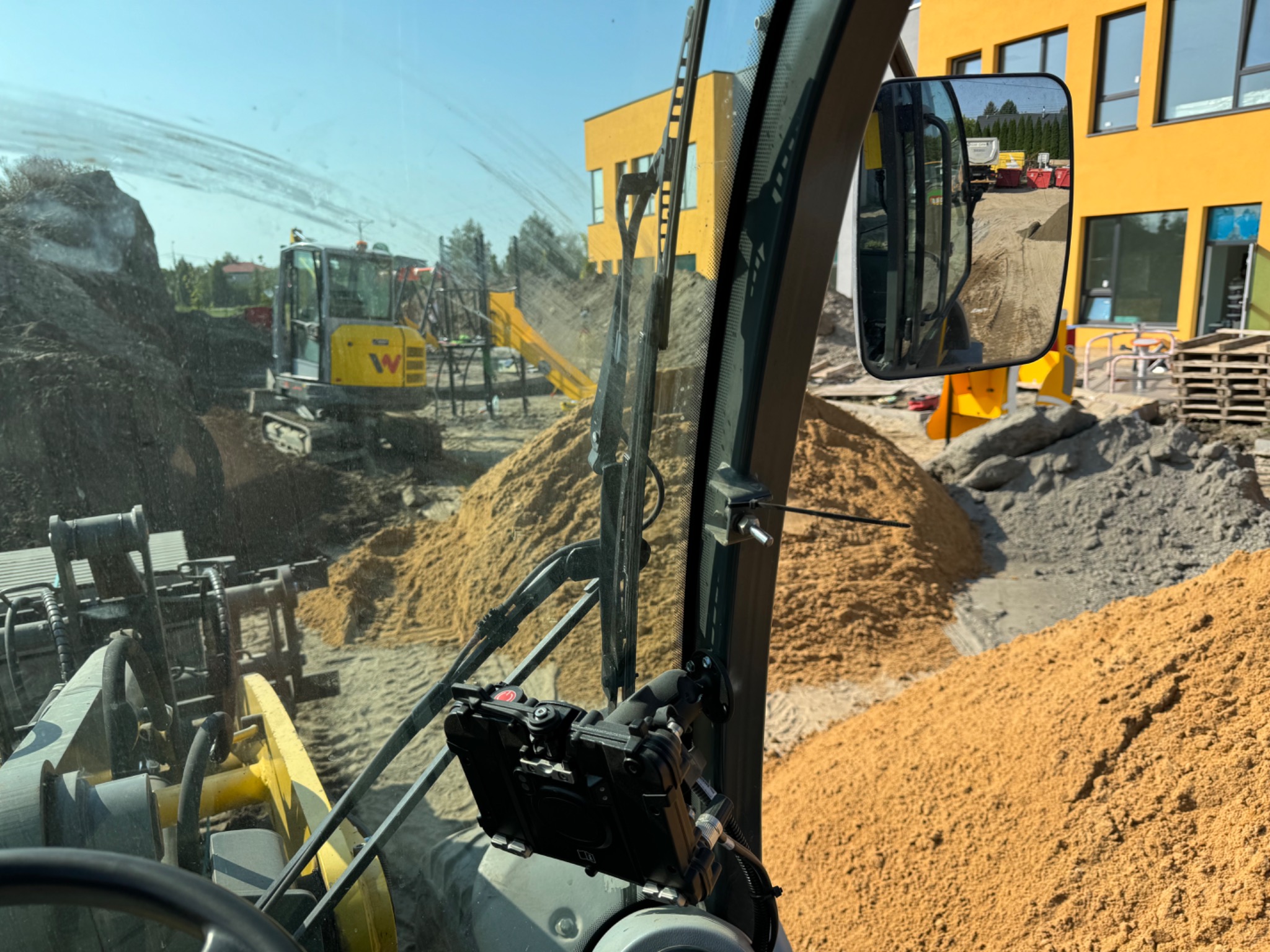 Widok z kabiny ładowarki na plac budowy z hałdami piasku i pracującą koparką w tle, z fragmentem żółtego budynku widocznym w lusterku.
