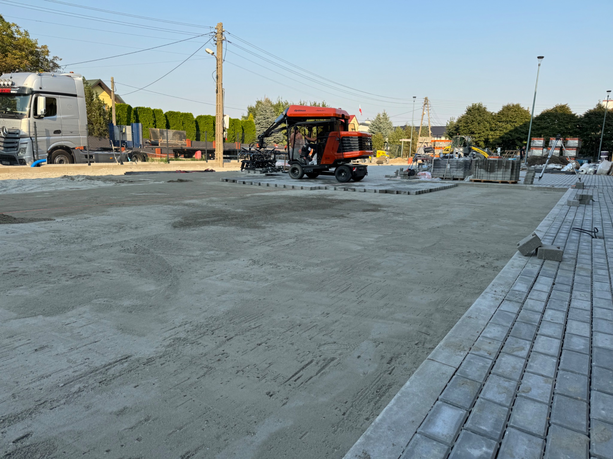 Plac budowy z przygotowanym podłożem, częściowo ułożoną szarą kostką brukową i pomarańczową maszyną do jej układania, widoczny również samochód ciężarowy.