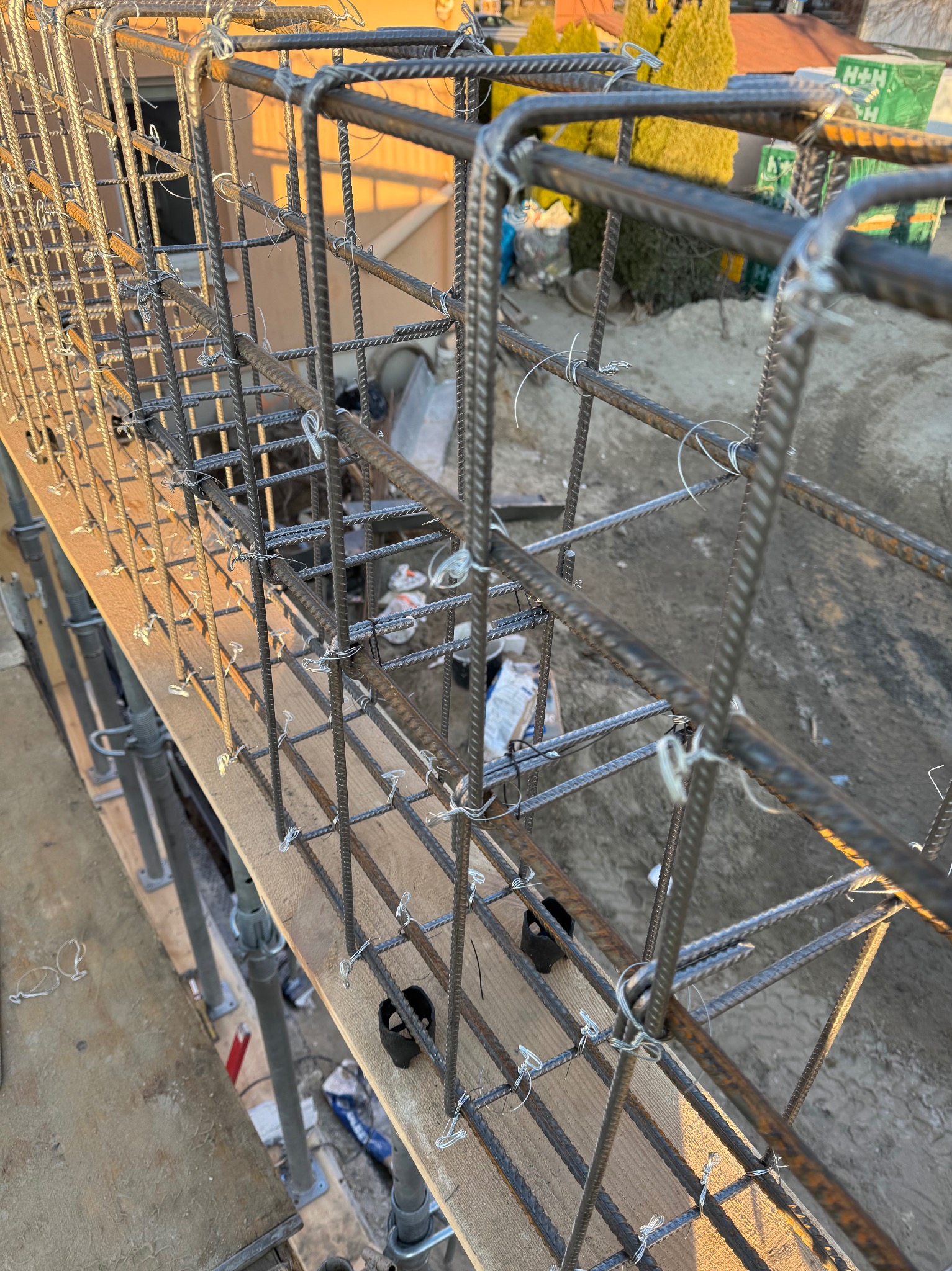 Zbrojenie fundamentu w formie stalowej klatki, połączone drutem wiązałkowym, wsparte na drewnianym szalunku i metalowych podporach, widoczne elementy krajobrazu w tle.