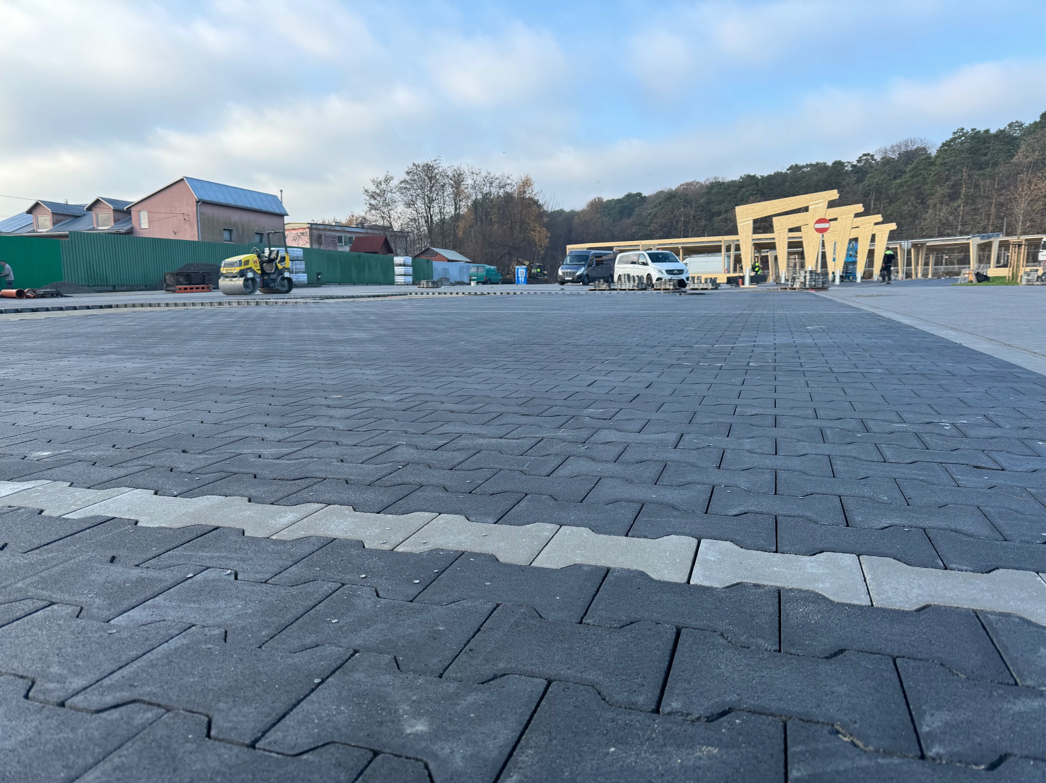 Rozległy plac wyłożony szarą kostką brukową z jasnoszarym pasem, w tle budynek w trakcie budowy, maszyna budowlana i zaparkowane samochody.