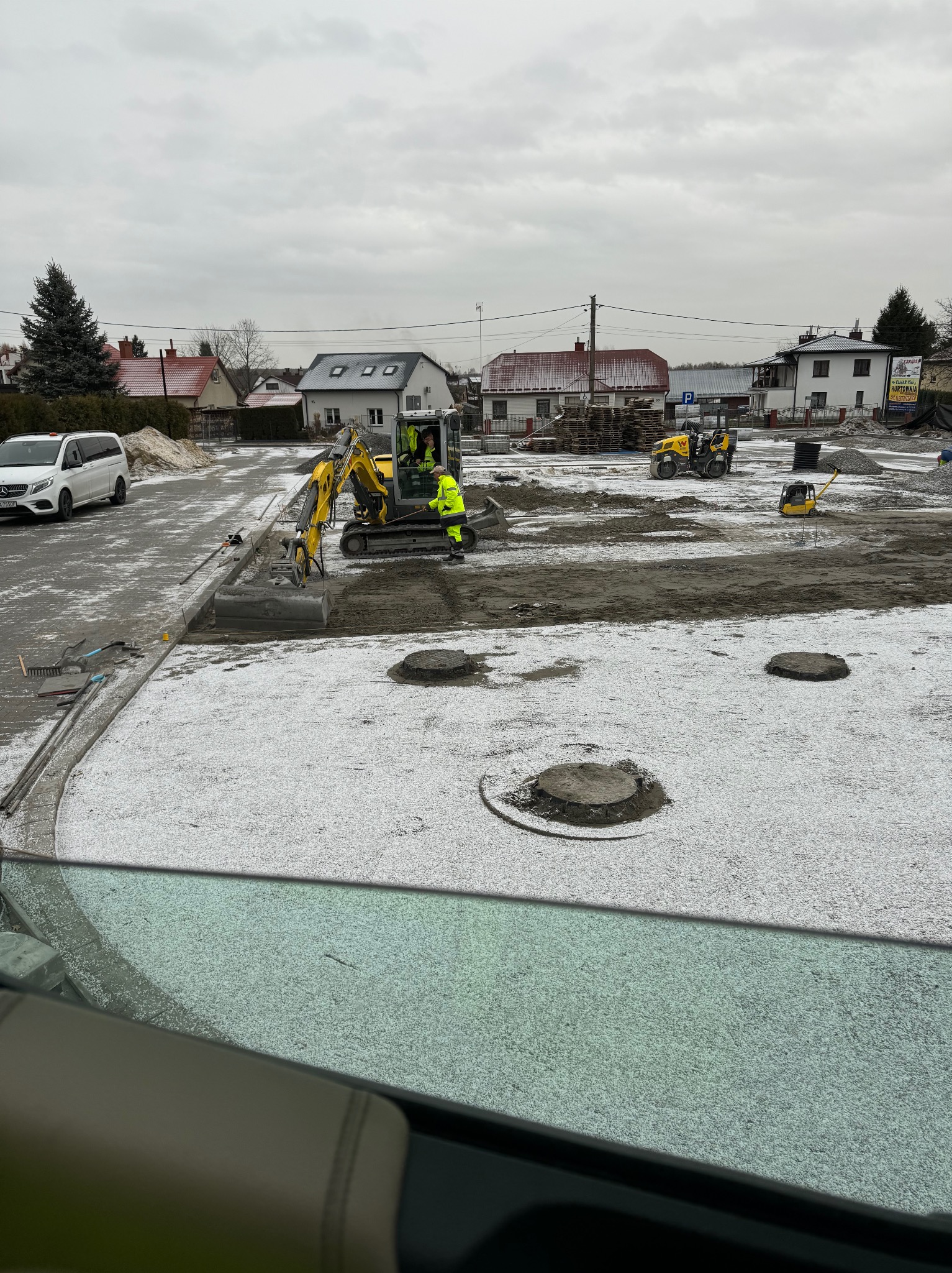 Żółta koparka z operatorem w kamizelce odblaskowej, przygotowuje teren pod chodnik z obrzeżami, w tle ubijarka i walec, całość lekko przyprószona śniegiem.