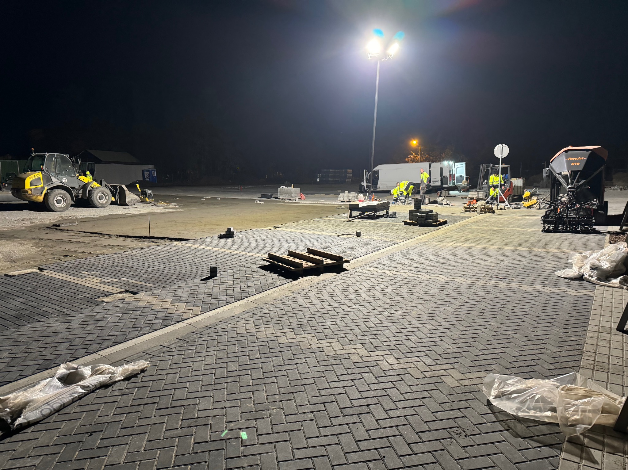 Nocna praca przy układaniu kostki brukowej na dużej powierzchni, widoczny sprzęt budowlany, paleta z kostką, ekipa w odblaskowych ubraniach i oświetlenie placu budowy.