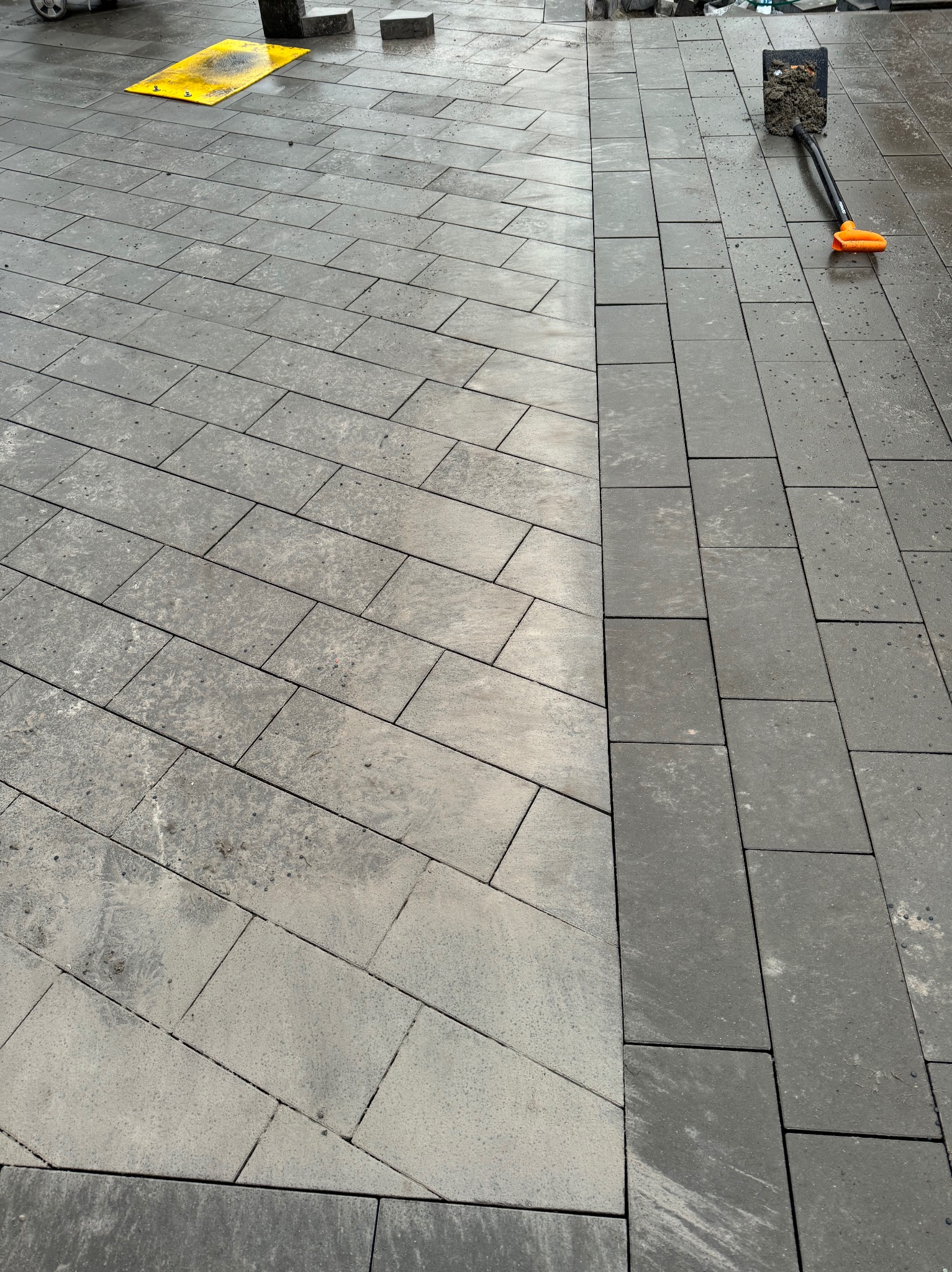 Ułożona część nawierzchni z szarej kostki brukowej w dwóch odcieniach, widoczna żółta mata ochronna i łopata z ziemią.