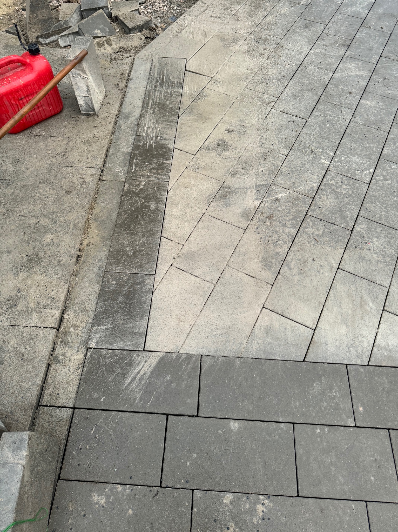 Fragment układanej nawierzchni z prostokątnych, szarych kostek brukowych, częściowo wilgotnych, z widocznym czerwonym kanistrem i łopatą w tle.