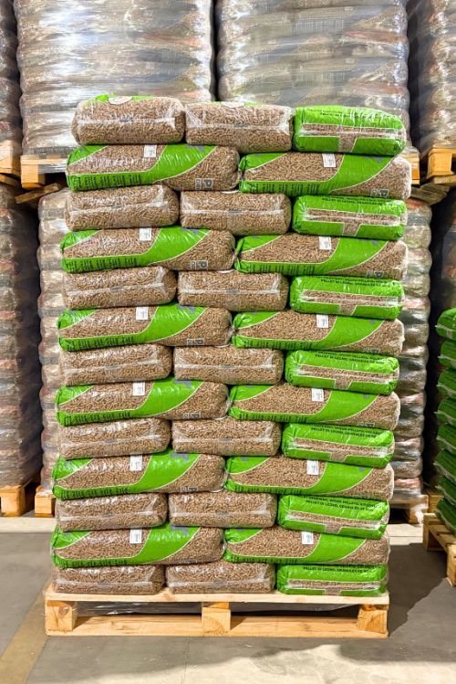 Paleta z ułożonymi workami pelletu drzewnego w folii, z zielonymi akcentami, magazynowana w hali. Widoczne zapasy pelletu w tle.