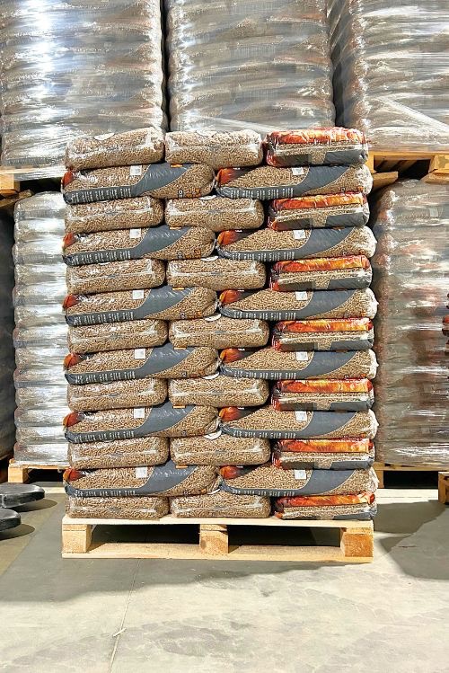 Stos worków z pelletem drzewnym ułożonych na palecie, widoczne także zapasy pelletu w przezroczystej folii. Magazyn opału.