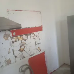 Ściana w kuchni w trakcie prac przygotowawczych do remontu, z fragmentami starej glazury i farby oraz widocznymi śladami po instalacjach.