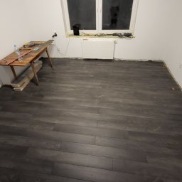 Iron development Łukasz Żelazko - Montaż paneli podłogowych w pokoju, widoczne narzędzia na stole i listwy przypodłogowe w trakcie instalacji, pomarańczowe kliny dystansowe przy ścianach.