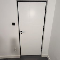 Iron development Łukasz Żelazko - Białe drzwi wewnętrzne z czarną ościeżnicą i klamką na tle białych ścian i ciemnej podłogi, widoczny czarny włącznik światła i gniazdko elektryczne.