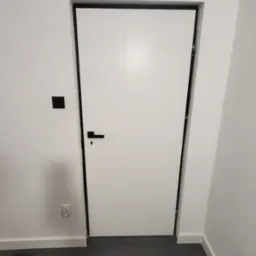 Białe drzwi wewnętrzne z czarną ościeżnicą i klamką na tle białych ścian i ciemnej podłogi, widoczny czarny włącznik światła i gniazdko elektryczne.