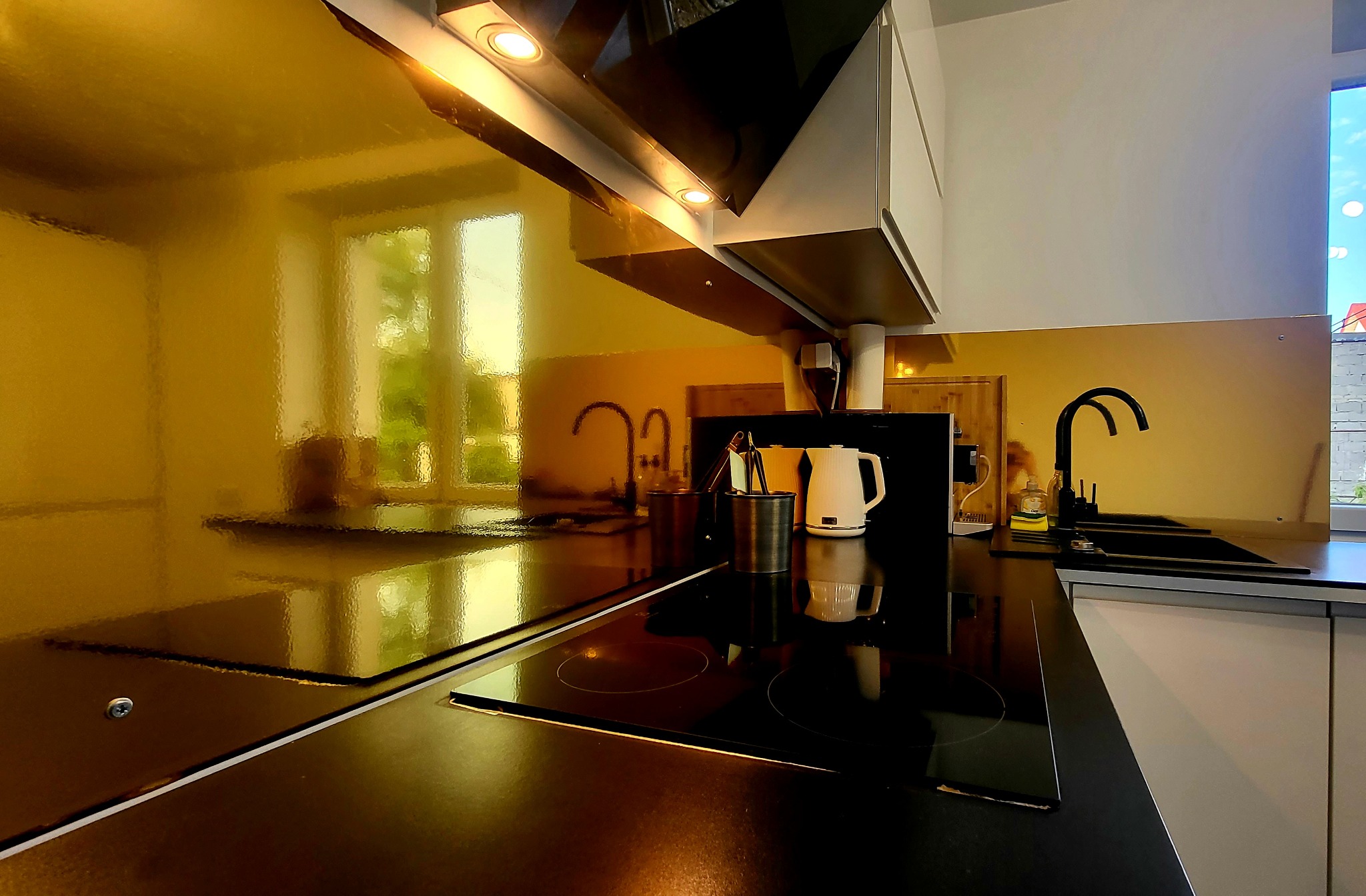 Nowoczesna kuchnia z czarnym blatem, złotą ścianą nad blatem odbijającą okno, indukcyjną płytą grzewczą i minimalistycznym okapem.