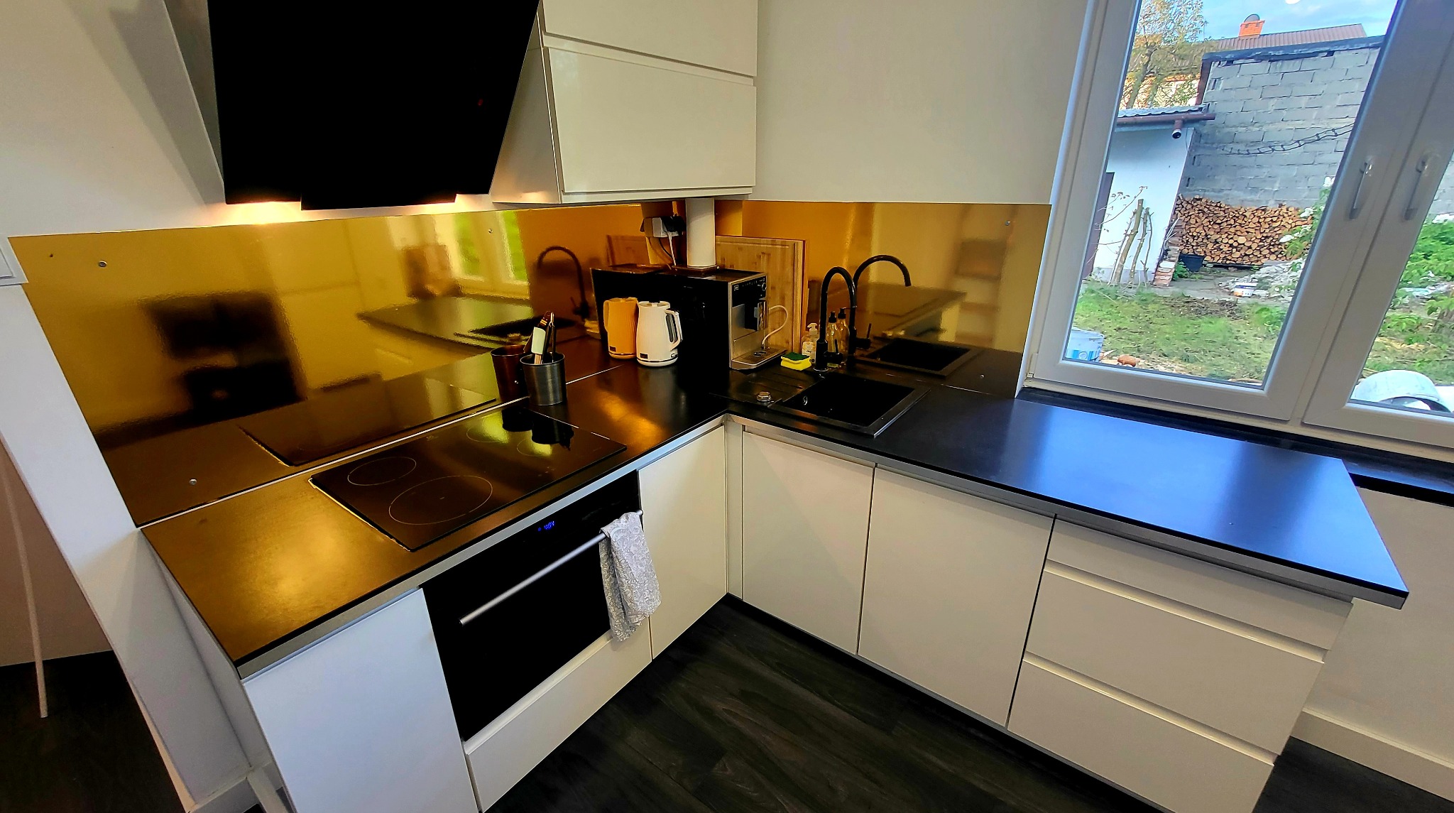 Nowoczesna kuchnia w odcieniach bieli i czerni, z złotym panelem nad blatem, widok na ogród przez okno, czarne blaty i sprzęt AGD, drewniana podłoga.