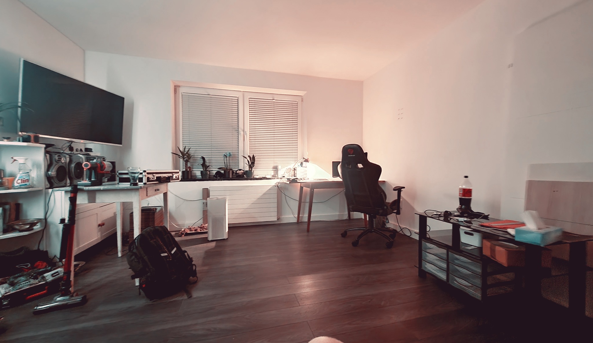 Pokój z biurkiem, krzesłem gamingowym, telewizorem, półkami z przedmiotami i plecakiem na podłodze.