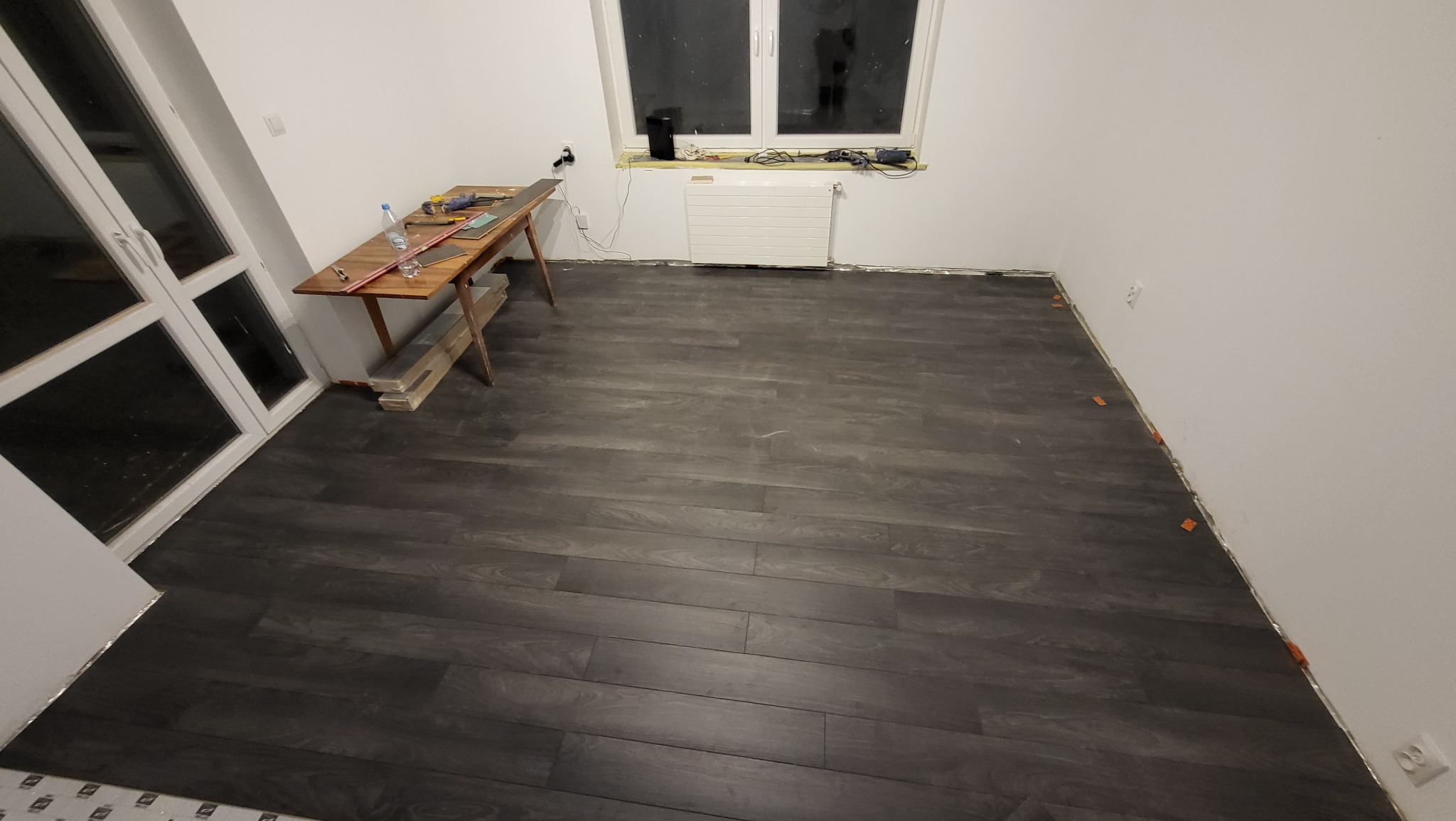 Montaż paneli podłogowych w pokoju, widoczne narzędzia na stole i listwy przypodłogowe w trakcie instalacji, pomarańczowe kliny dystansowe przy ścianach.