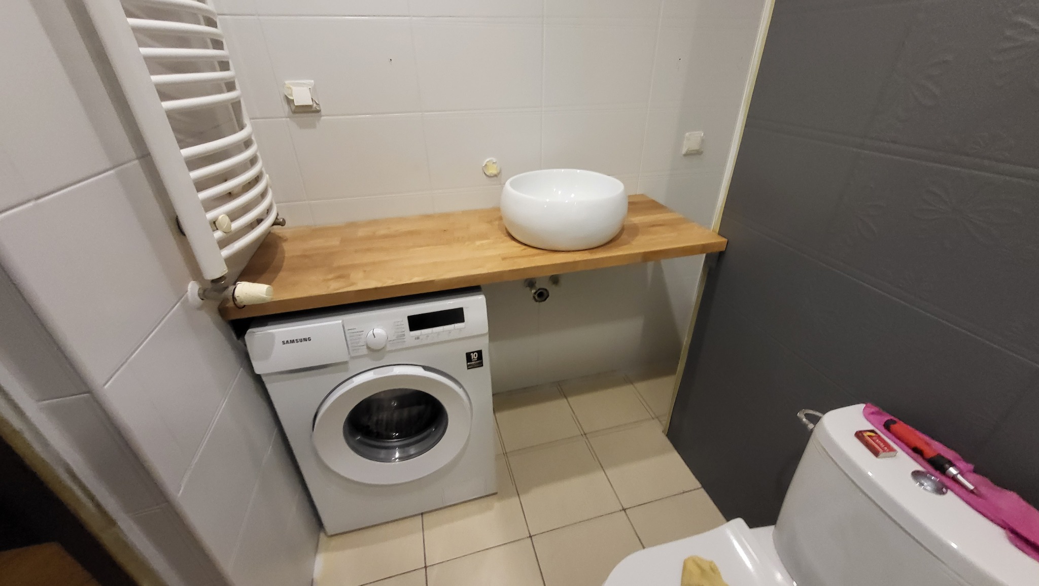 Biała umywalka nablatowa na blacie z jasnego drewna nad pralką w małej łazience z grzejnikiem drabinkowym i szarą ścianą z dekoracyjnymi panelami.