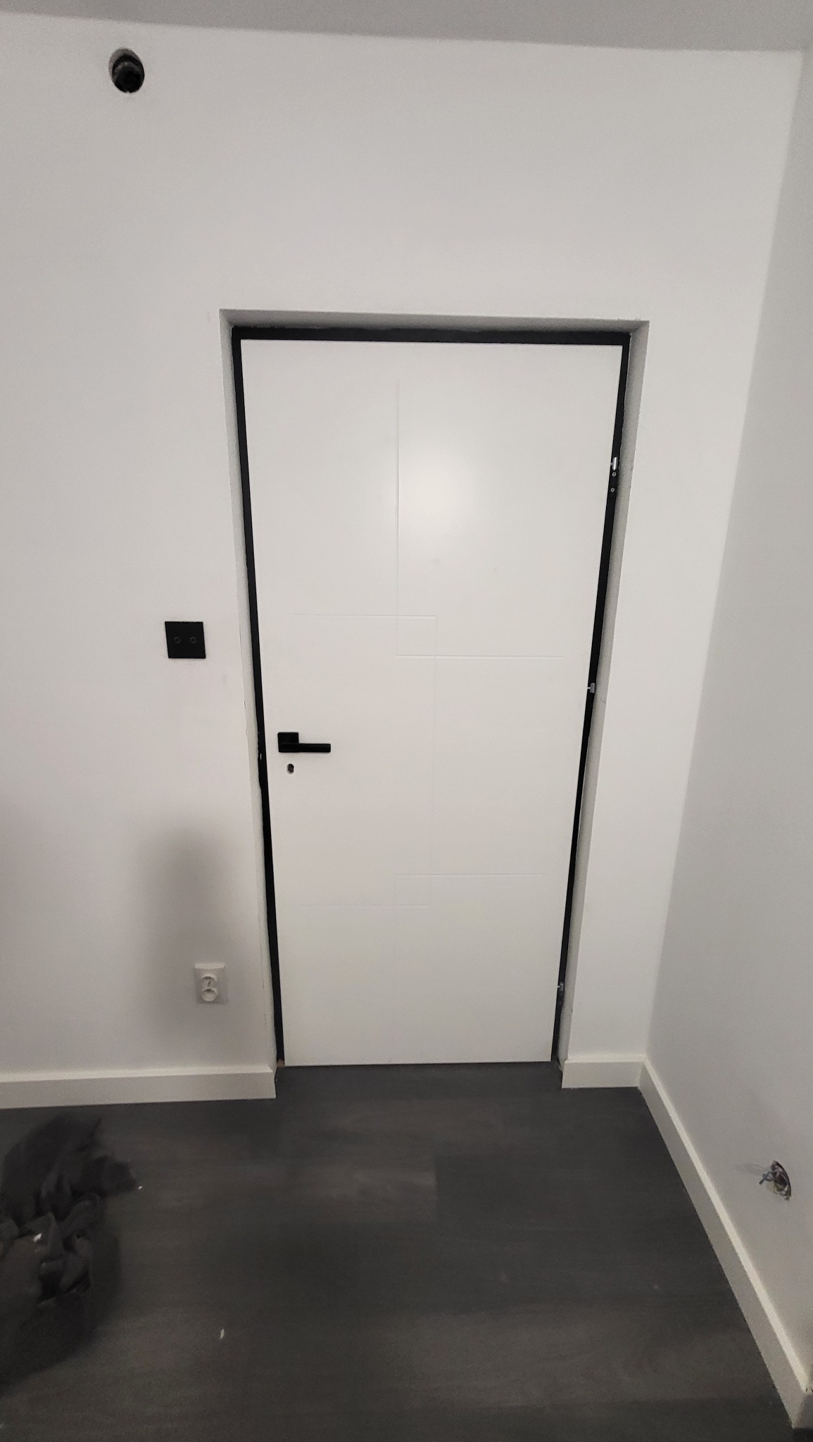 Białe drzwi wewnętrzne z czarną ościeżnicą i klamką na tle białych ścian i ciemnej podłogi, widoczny czarny włącznik światła i gniazdko elektryczne.