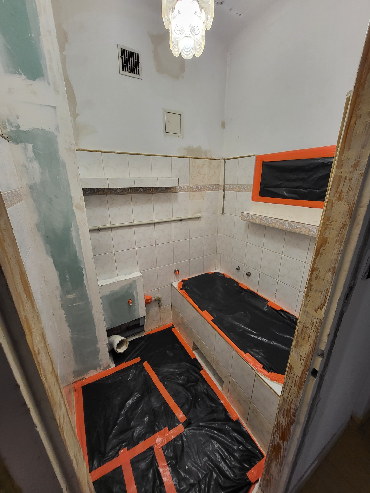 Łazienka w trakcie remontu: zabezpieczona folią i taśmą podłoga, ściany z częściowo położonymi płytkami, przygotowane do malowania, widoczne niedokończone prace przy instalacji wodnej i oświetleniu.