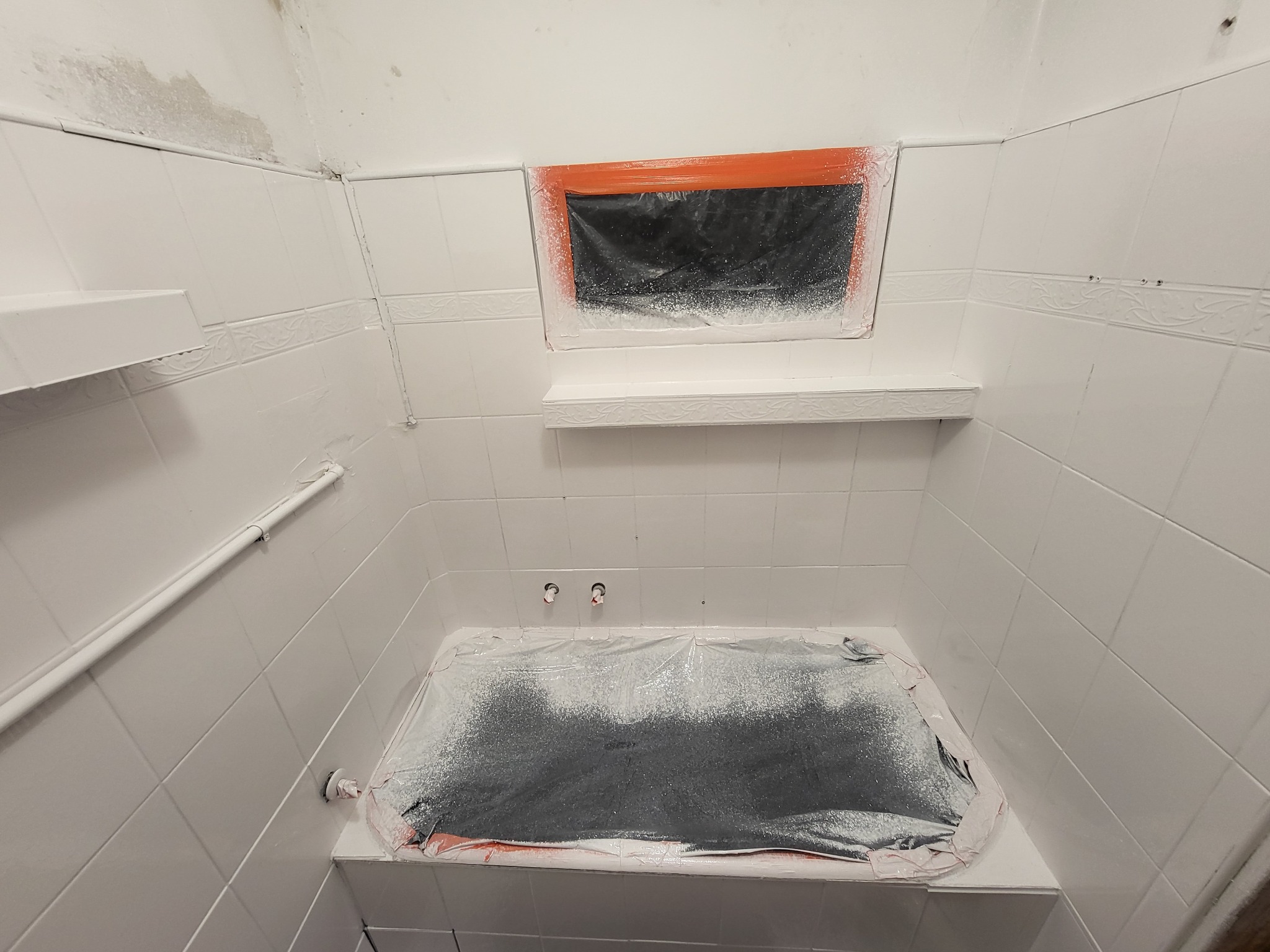 Przygotowanie łazienki do malowania: zakryta folią wanna, oklejone okno, białe płytki częściowo pomalowane, ślady farby na fugach i armaturze.