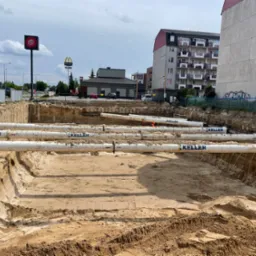 Głęboki wykop w piaszczystej ziemi z ułożonymi białymi rurami z logo Keller, w tle budynki mieszkalne i znaki Pizza Hut oraz McDonald's.