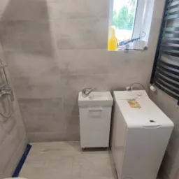 Jasna łazienka po remoncie: szare płytki, biała umywalka z szafką, pralka i czarny grzejnik drabinkowy. Nowoczesny design, schludne wykończenie.