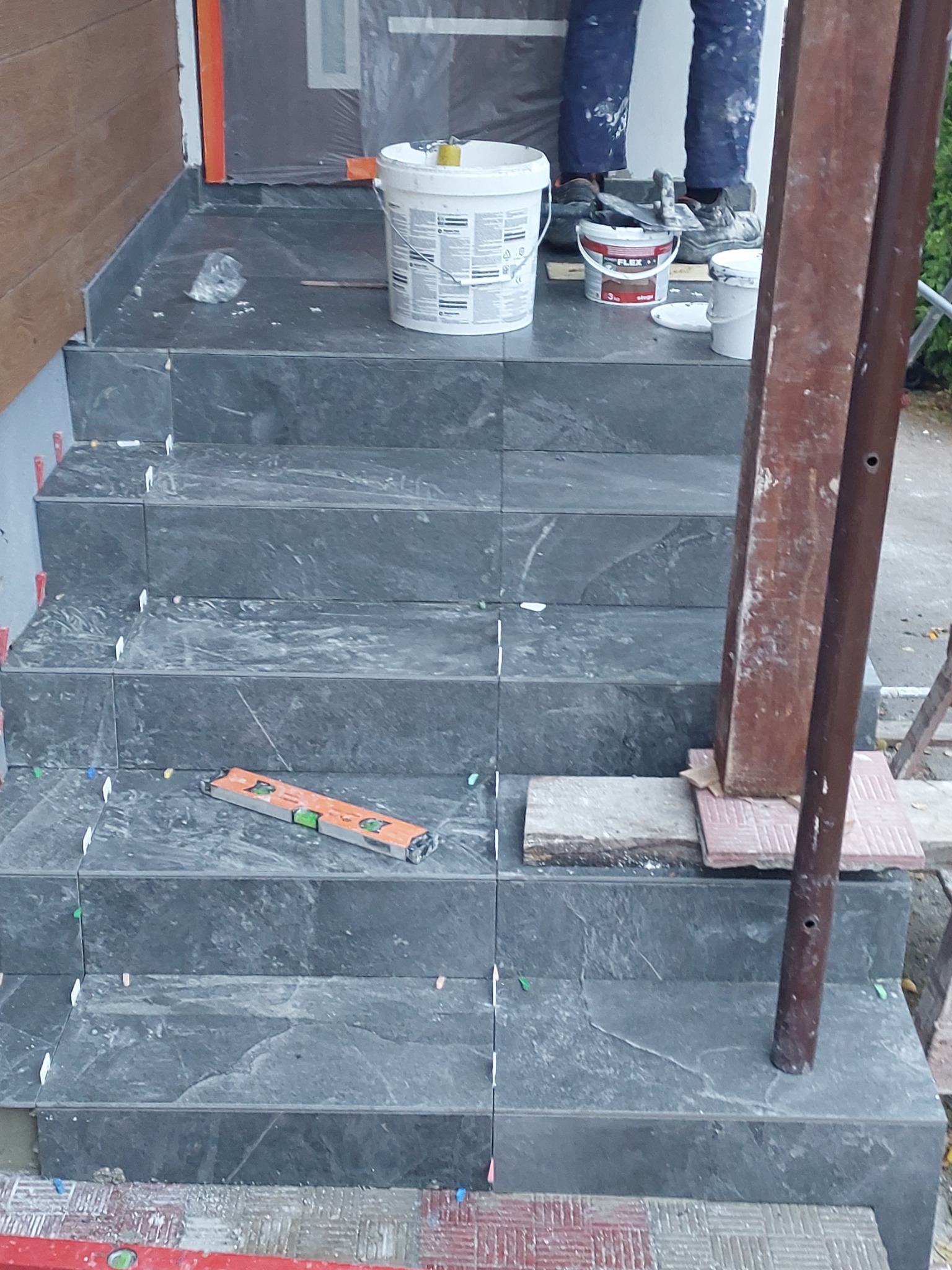 Układanie płytek na schodach wejściowych, widoczne poziomica, wiadro z klejem, krzyżyki dystansowe i ubrudzony klejem pracownik.